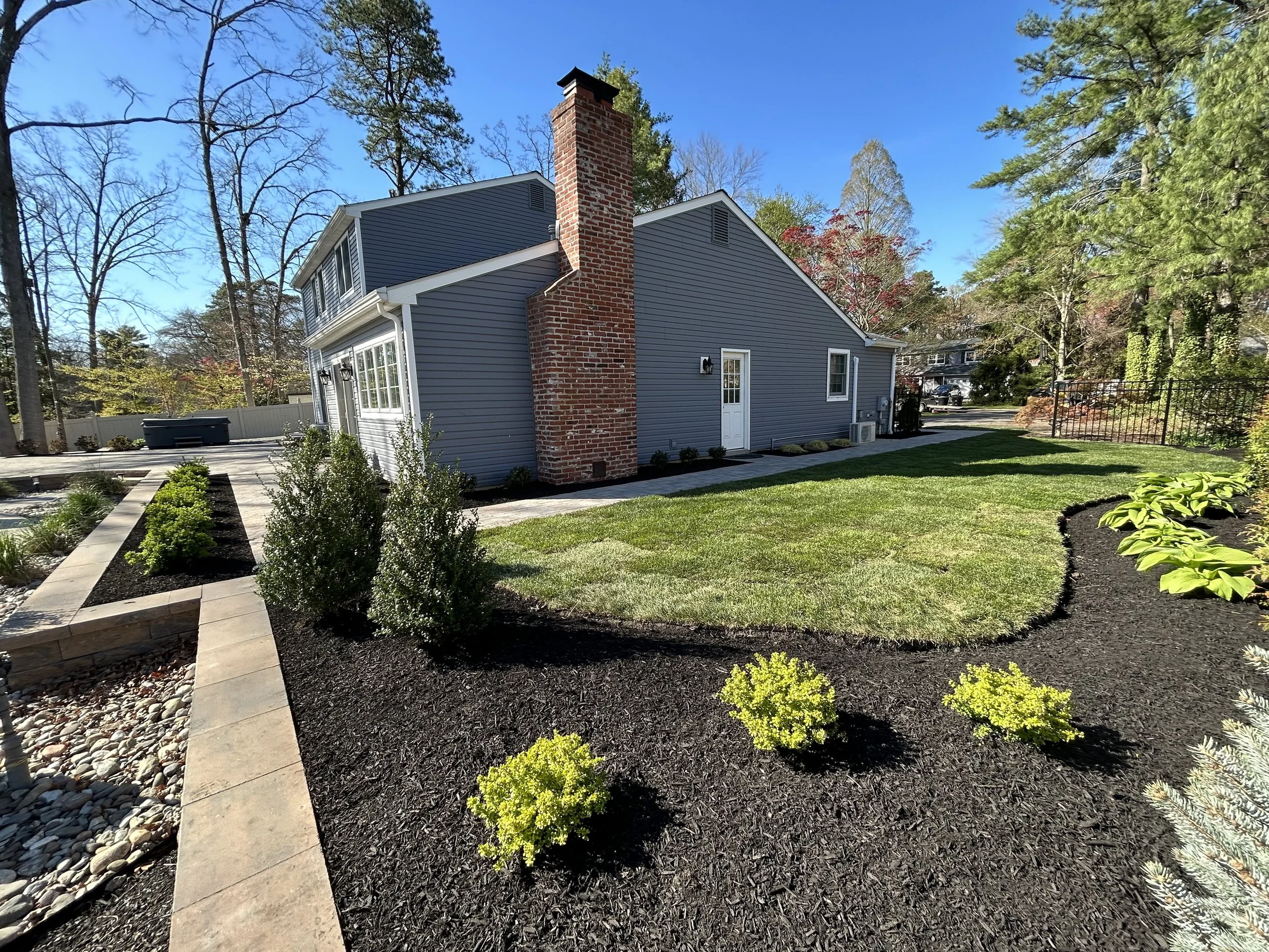Backyard yard with a house, a large brick chimney, and well-maintained lawn and garden with shrubs and plants, under a clear blue sky.