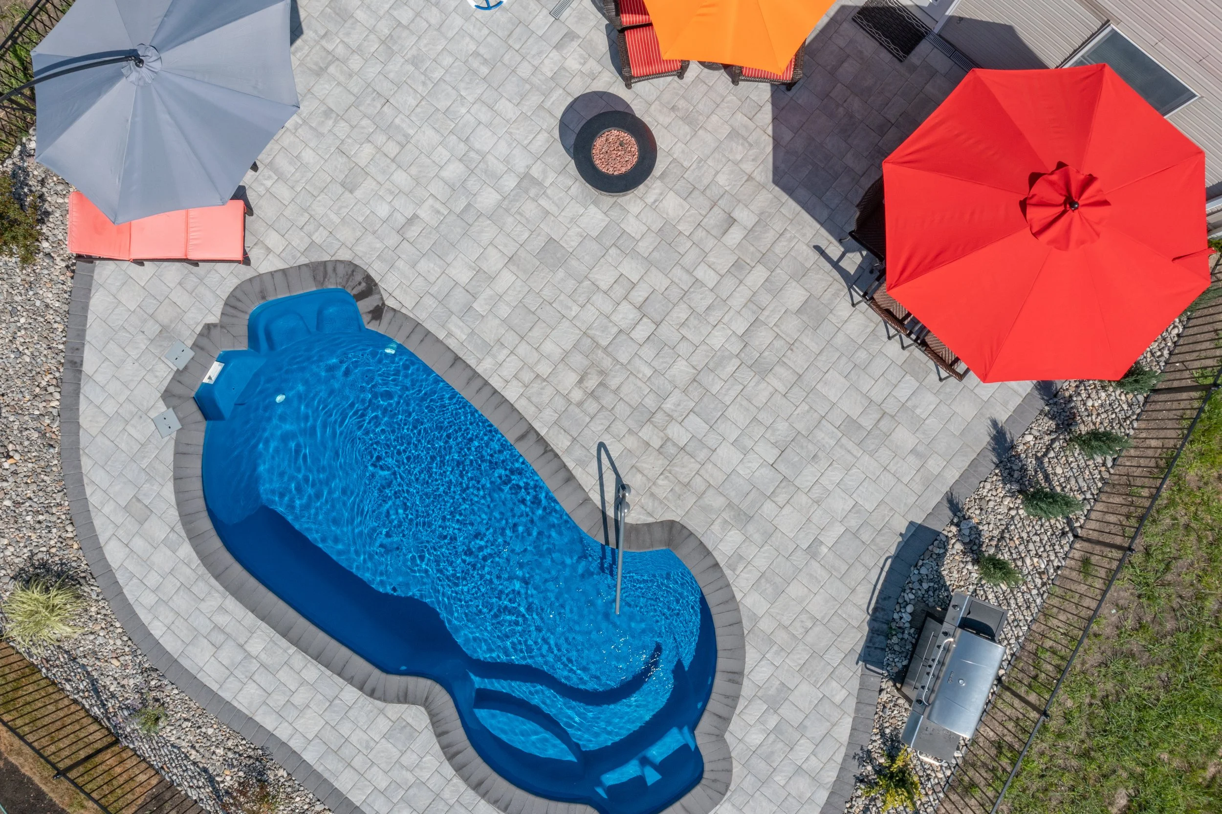 Aerial view of a backyard patio with a kidney-shaped swimming pool, a large red umbrella, a gray umbrella, and outdoor furniture, including chairs and a table, with a grill and landscaping.