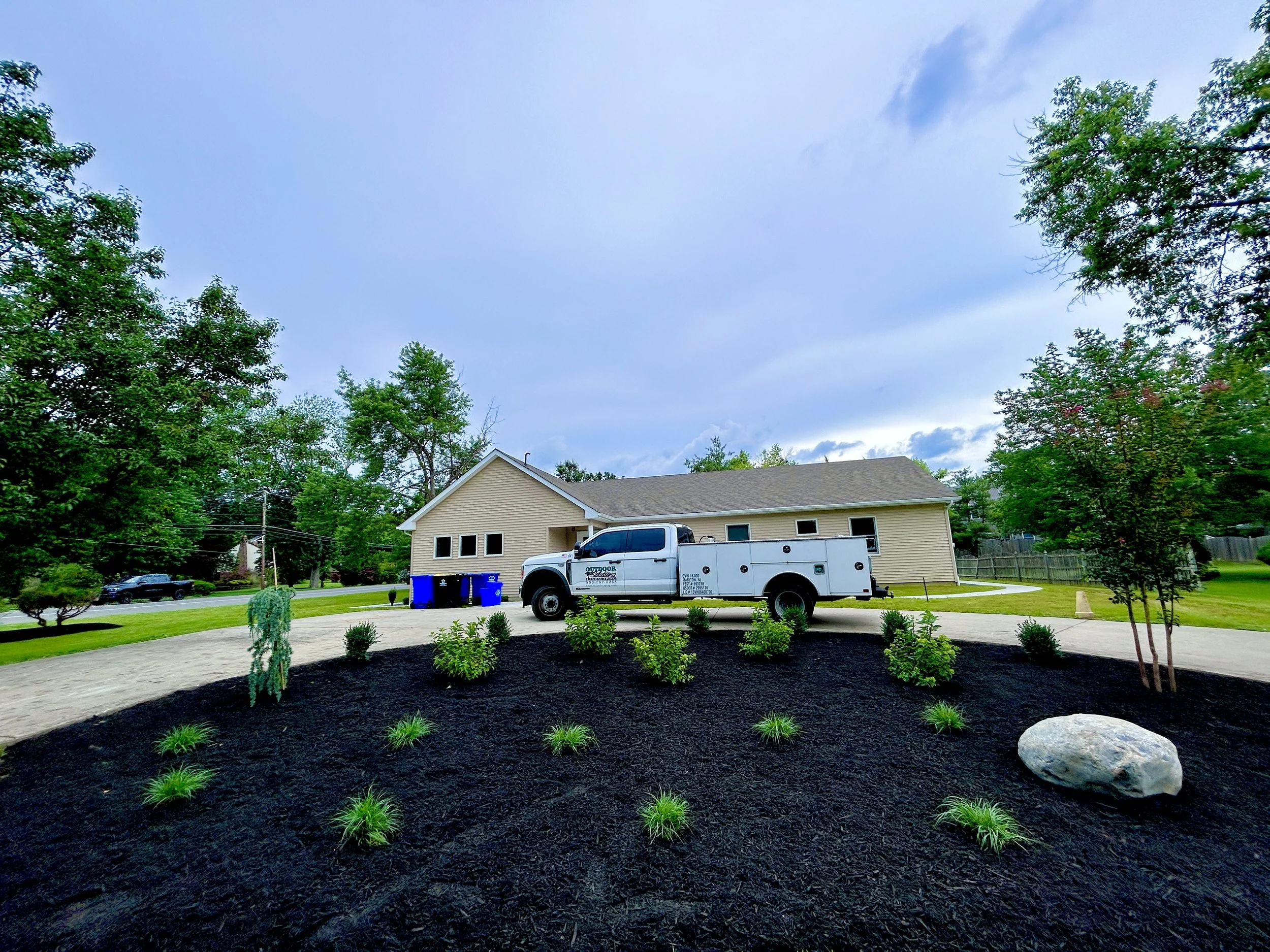 Residential house with landscaped yard featuring shrubs, a large rock, and a white utility truck parked in the driveway during daytime.