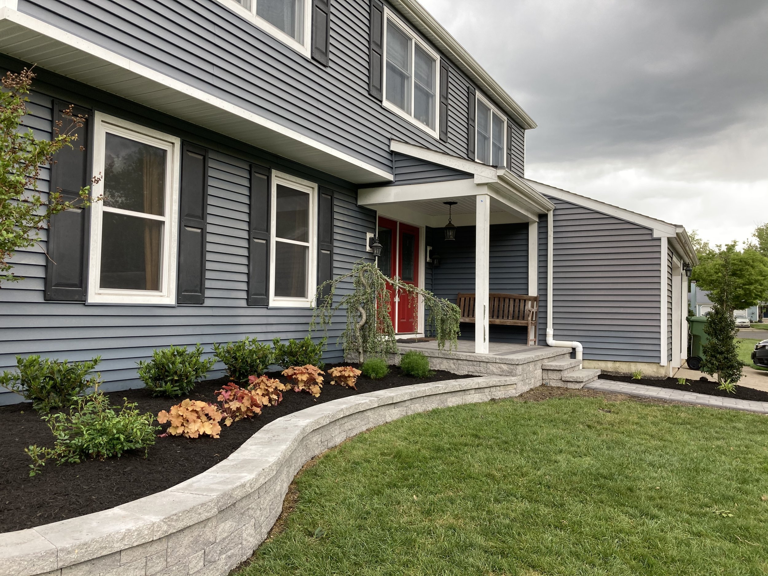 Front view of a modern house with blue siding, black shutters, a porch with a bench, and a landscaped yard with mulch, bushes, and a curved stone border