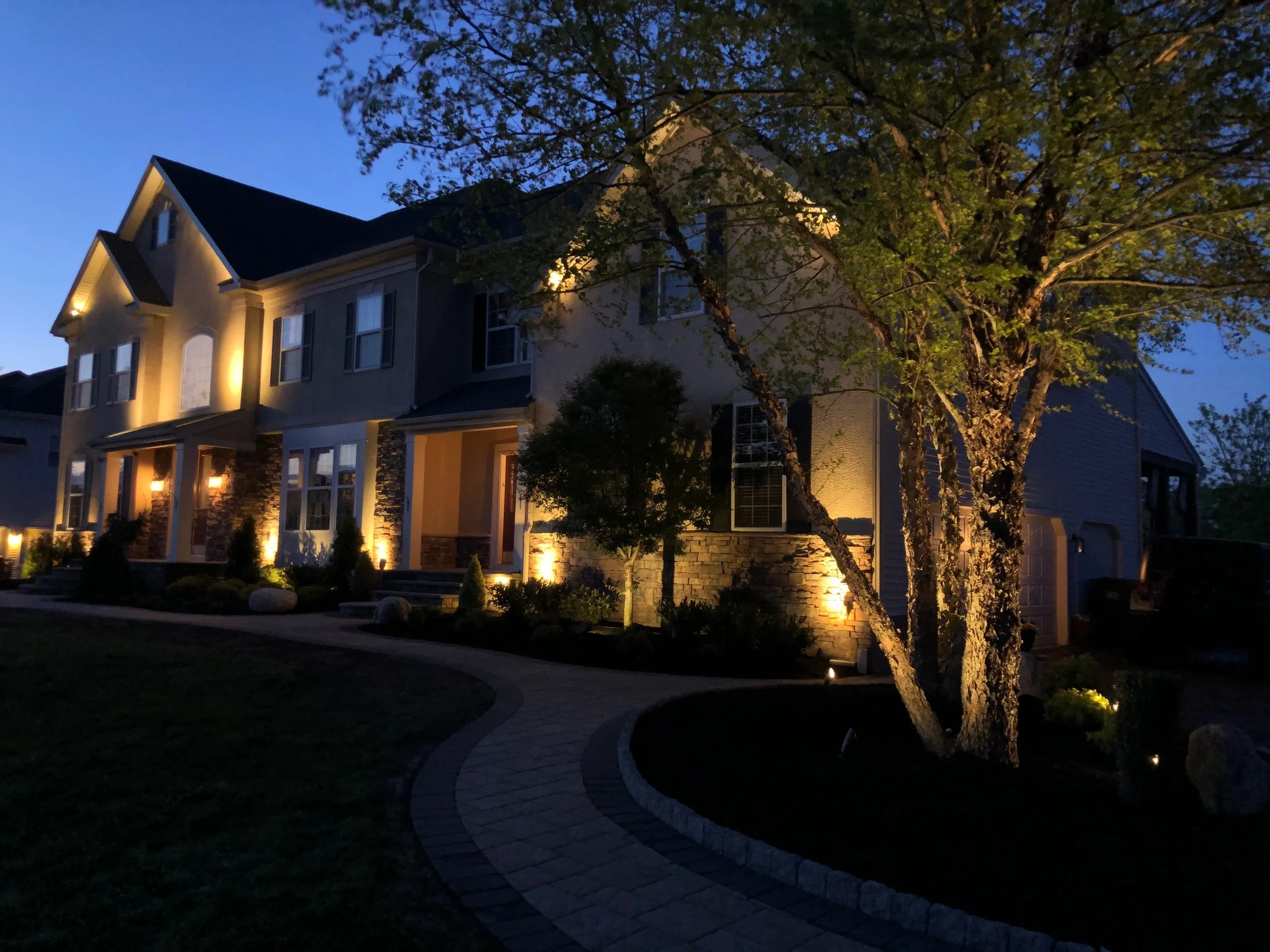 Nighttime view of a modern two-story house illuminated with exterior lighting, surrounded by landscaped garden and a curved stone walkway, with trees partly obscuring the house.
