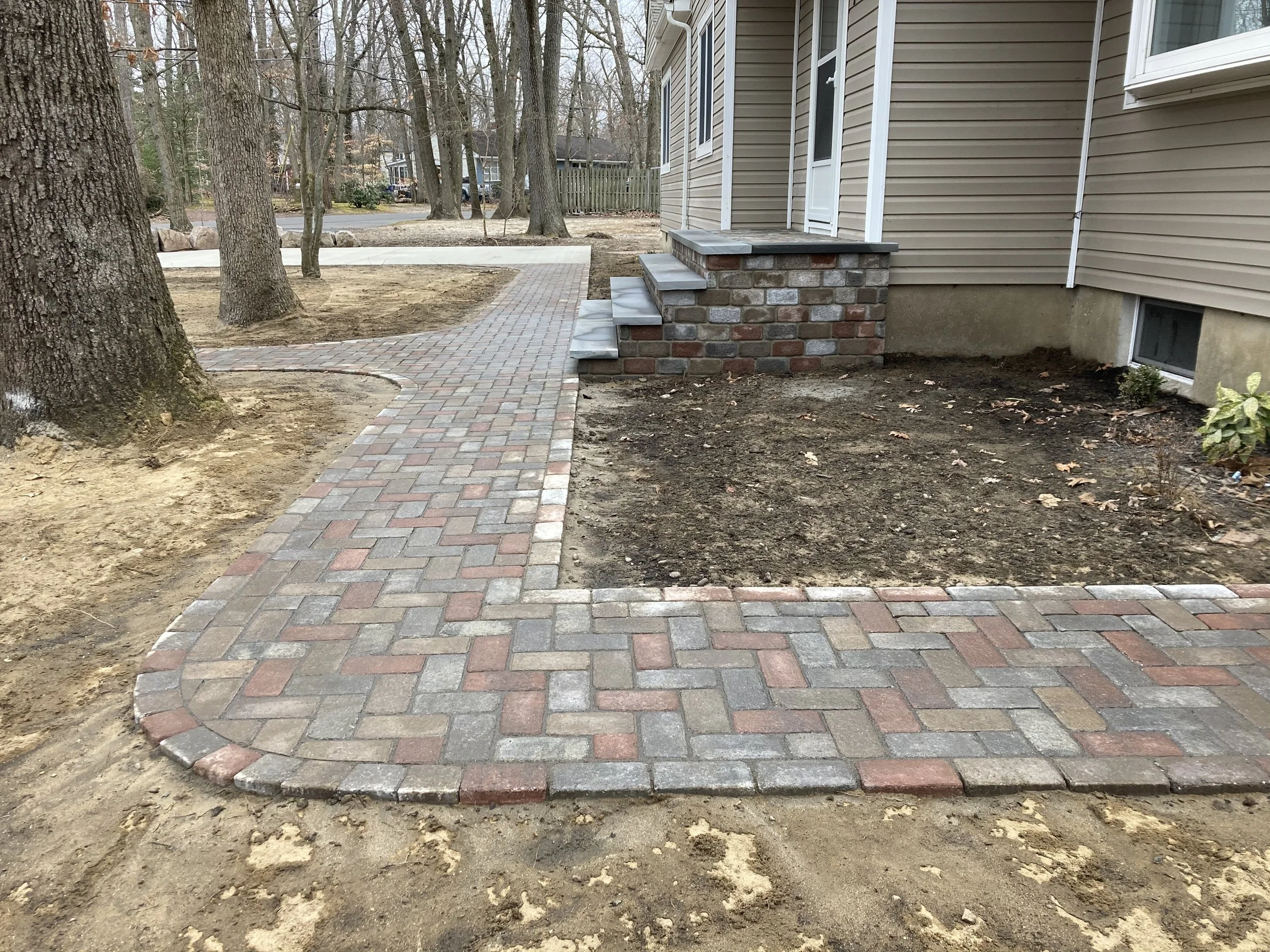 Newly completed brick sidewalk with stairs leading to the house entrance, surrounding trees, and patches of bare soil in the yard.