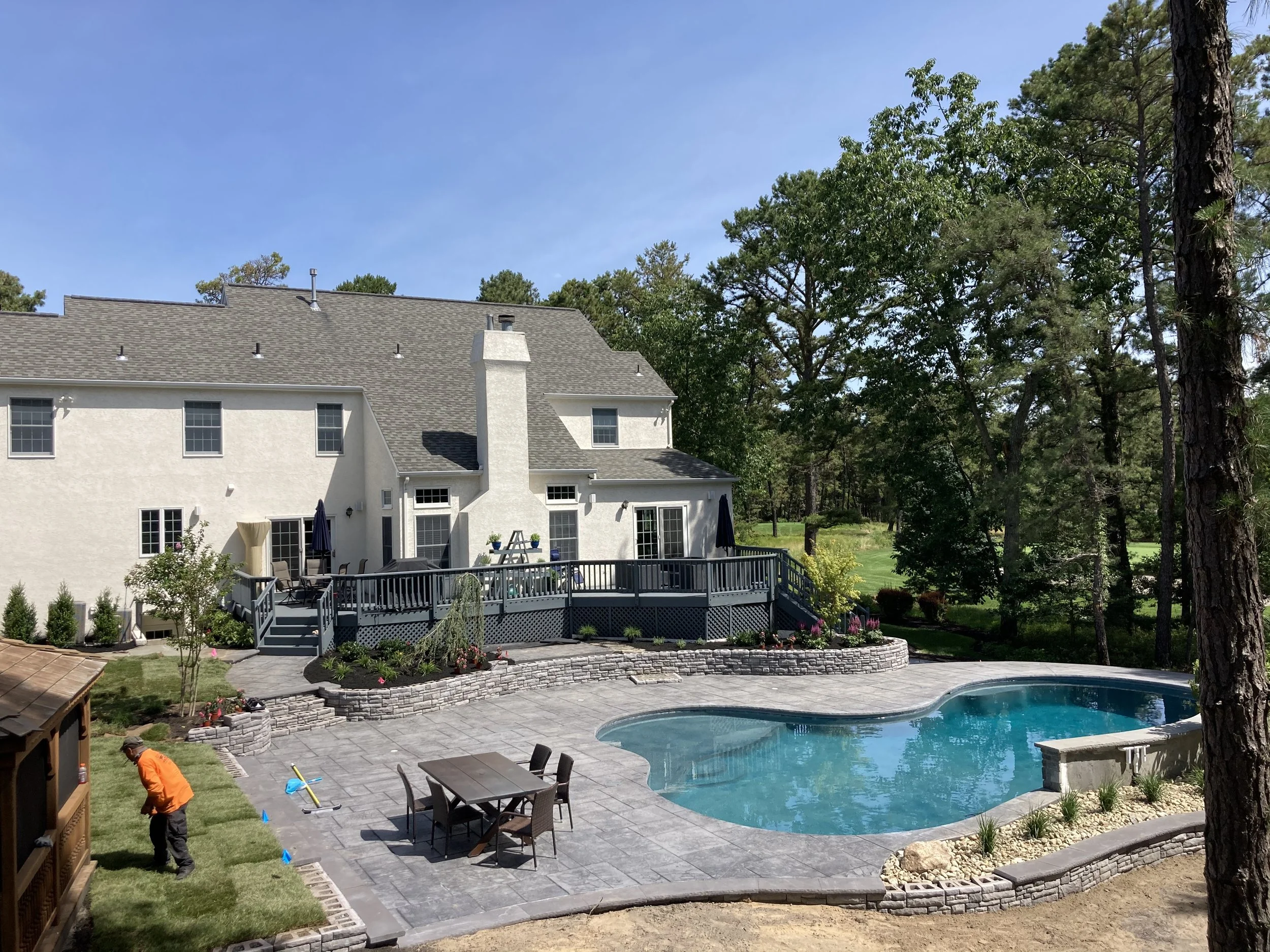Backyard with swimming pool, patio area with table and chairs, house in the background, person in orange shirt working on the grass, surrounded by trees and greenery under a blue sky.