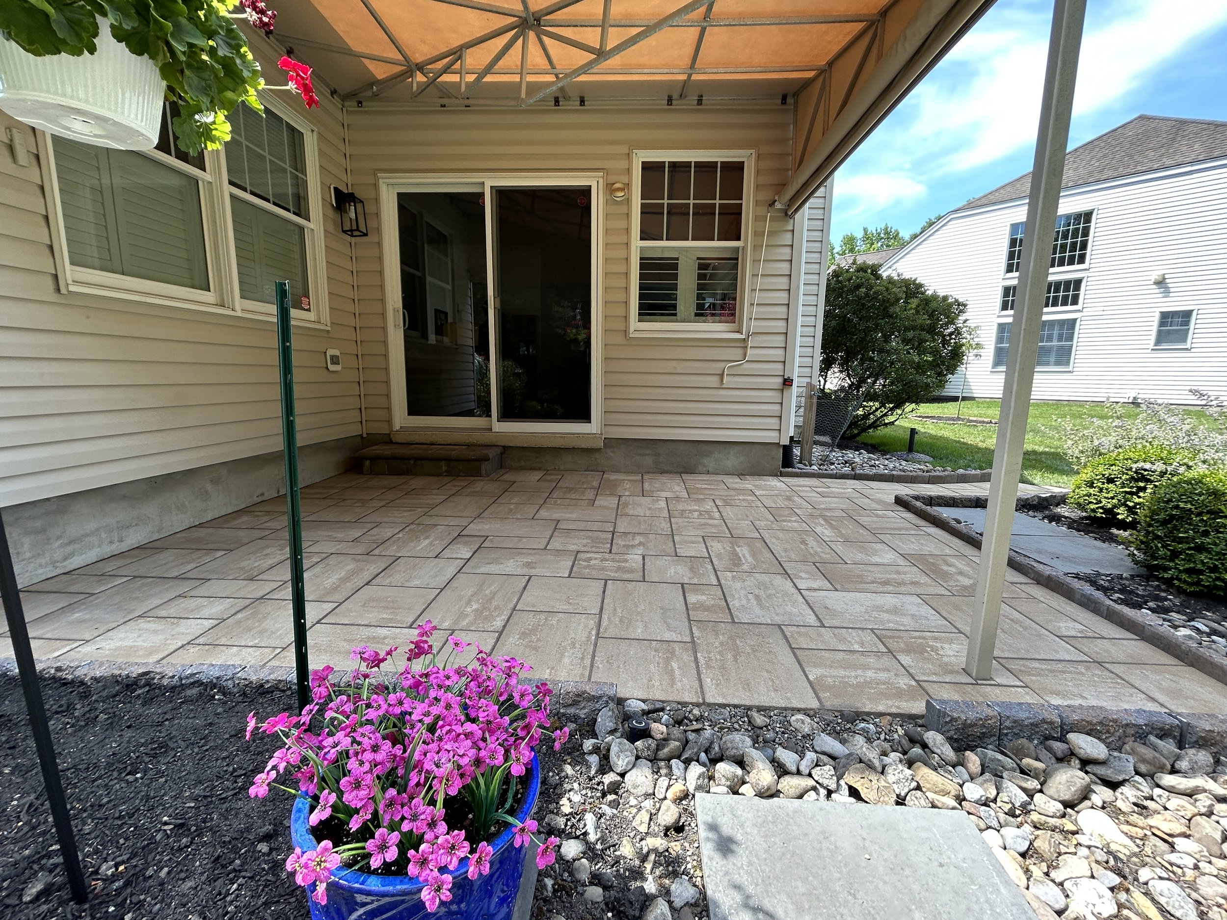 Backyard patio with beige stone tiles, potted pink flowers, garden beds, and a beige house with sliding glass door and windows.