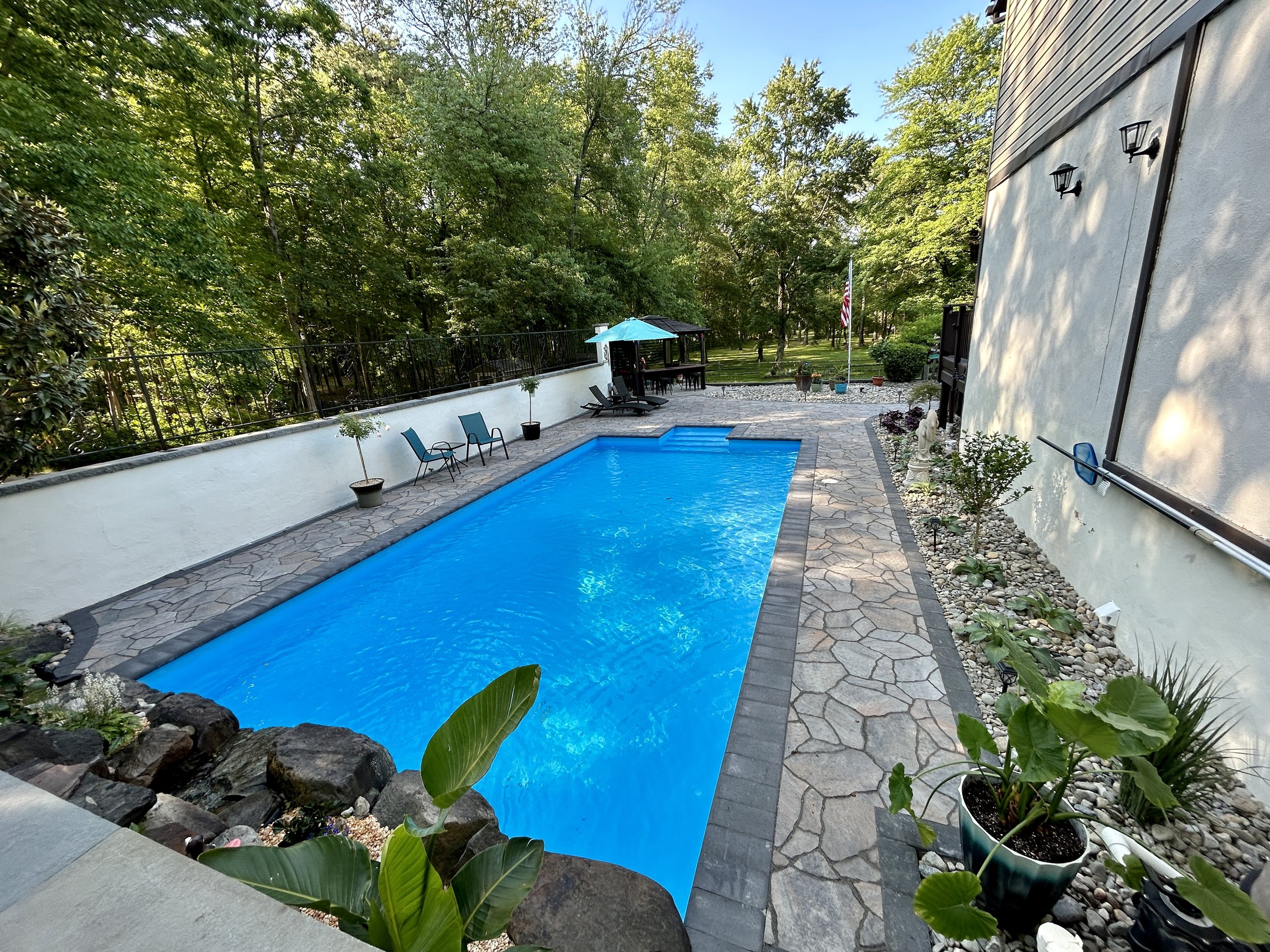 Backyard with a rectangular swimming pool, potted plants, stone pathway, outdoor chairs, and a gazebo with umbrella, surrounded by trees and fence.