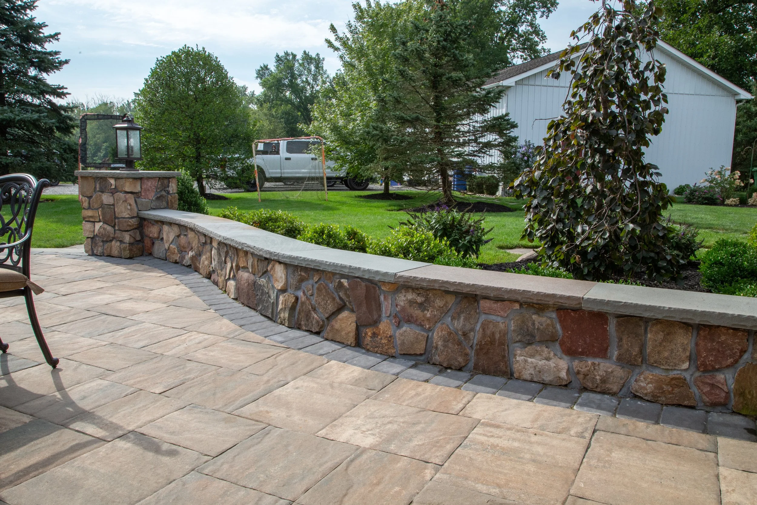 A backyard patio with a stone and concrete retaining wall, wrought iron chairs, and a view of a well-maintained lawn with trees, a shed, and a small soccer goal.