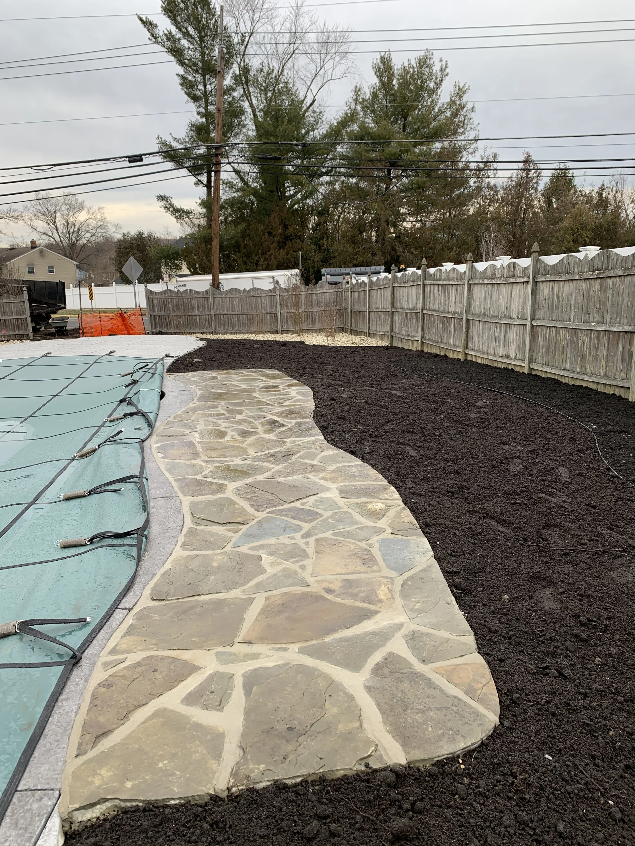 A backyard with a stone walkway, a covered swimming pool on the left, and a wooden fence enclosing the yard. The ground has dark soil, and there are trees and power lines in the background.