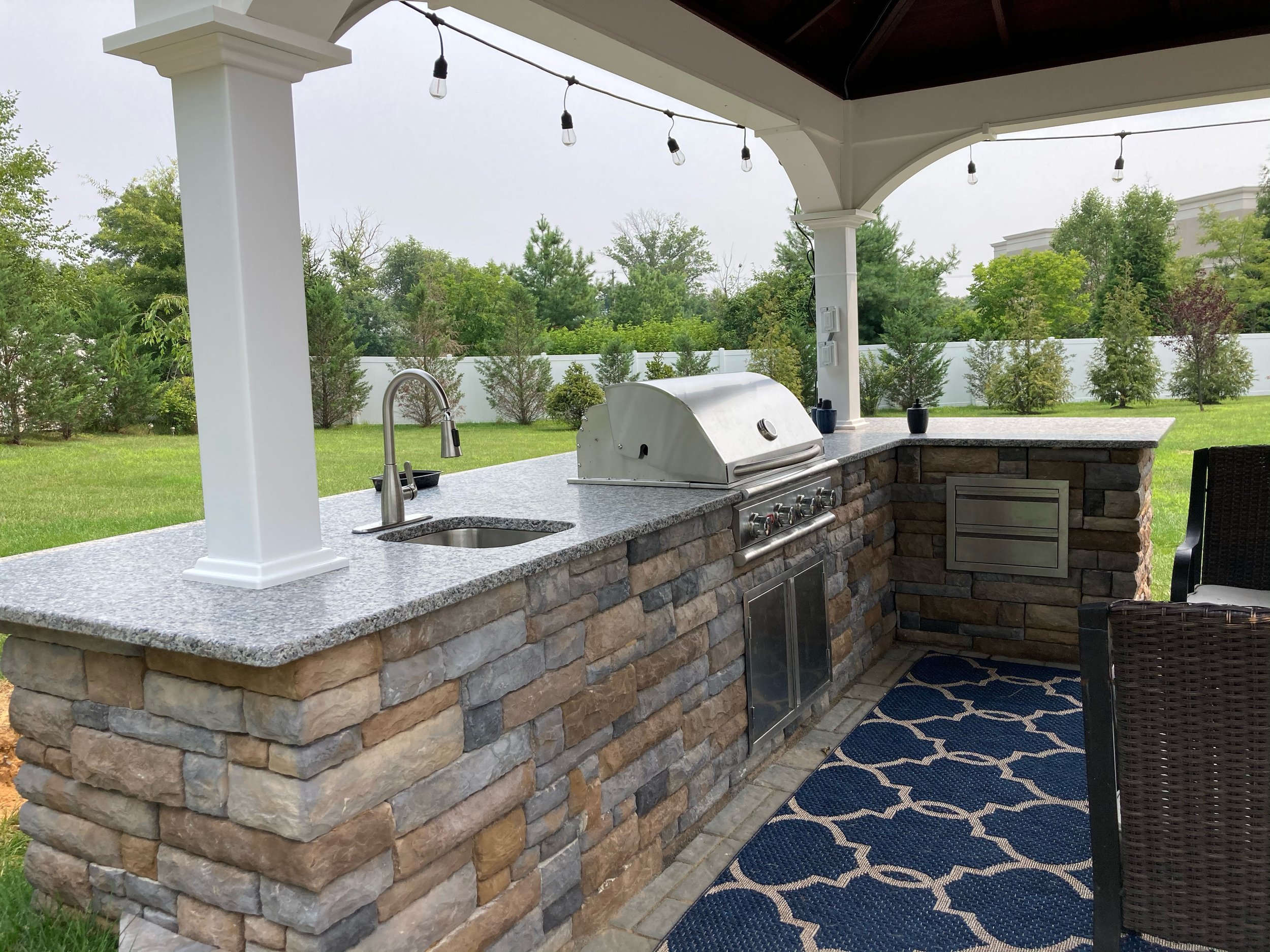 Outdoor kitchen with stone counter, built-in grill, sink, and decor items, under a covered patio with string lights, overlooking a backyard with green grass and trees.