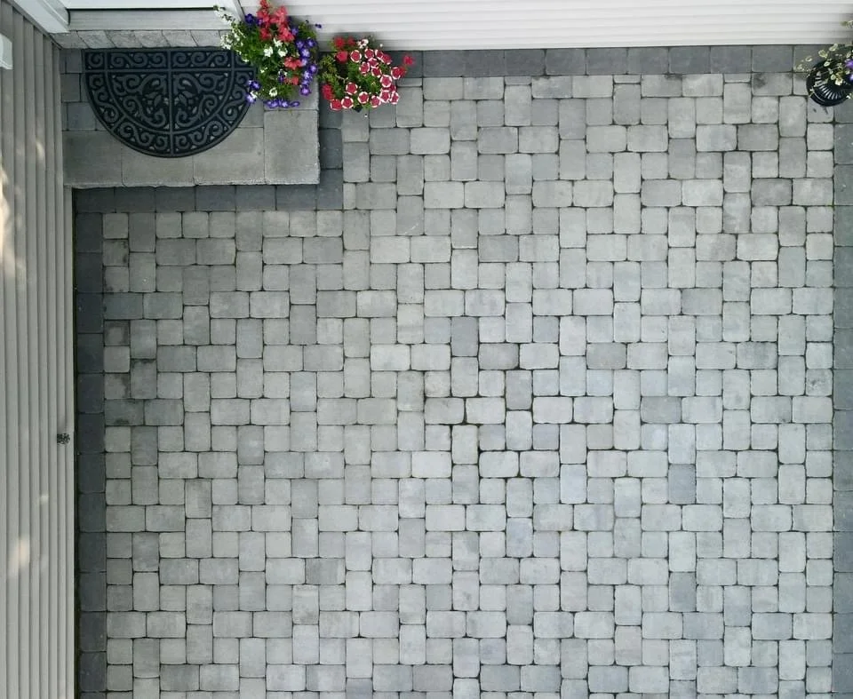 Top-down view of a paved outdoor area with gray cobblestone bricks. In the upper left corner, there are colorful potted flowers next to a black metal door mat. The upper right corner has a small potted plant. The bottom left has the edge of a corruga