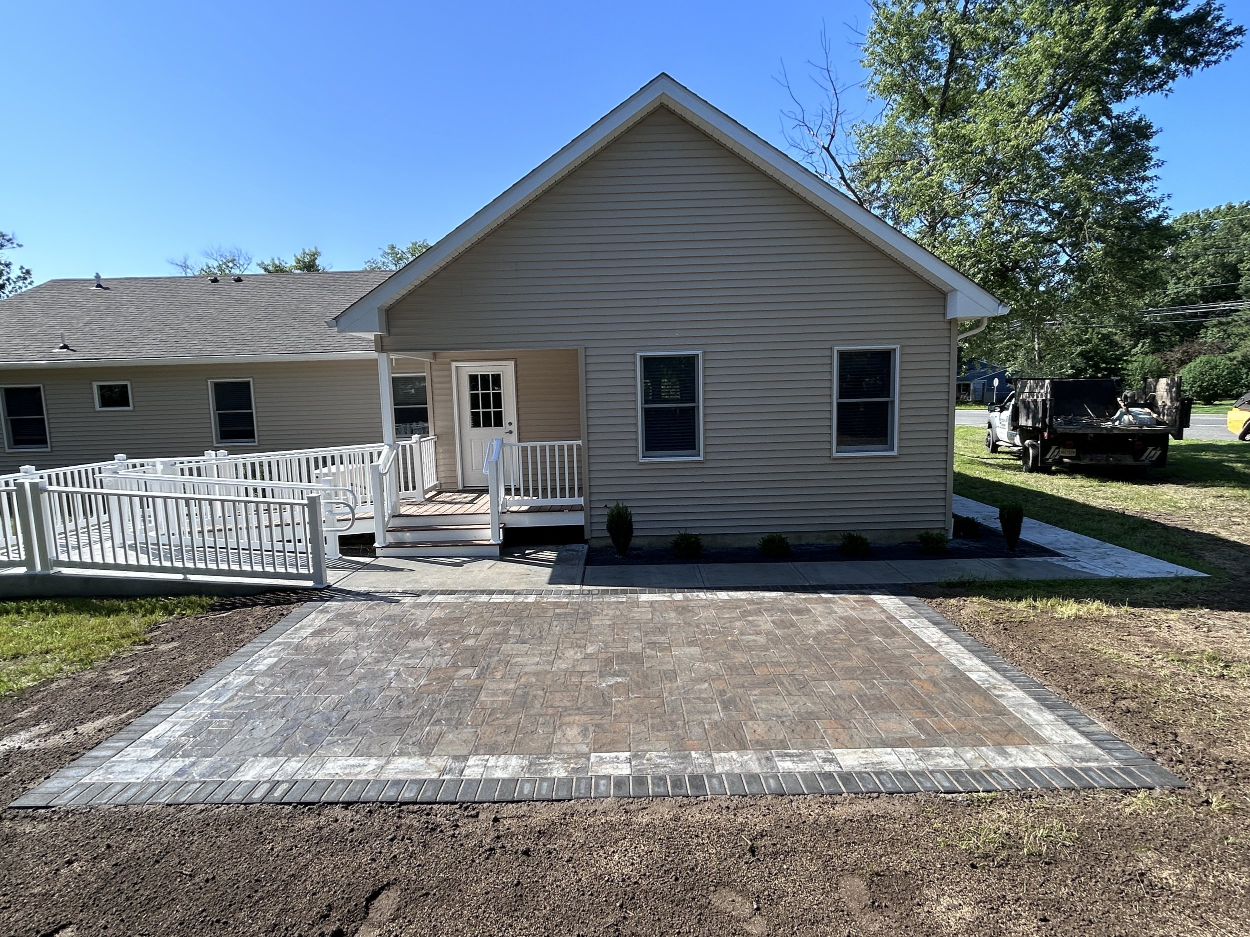 View of the backyard of a house with a new paved patio, a white ramp leading to a porch, and a white door with windows, beige siding, and a gabled roof.