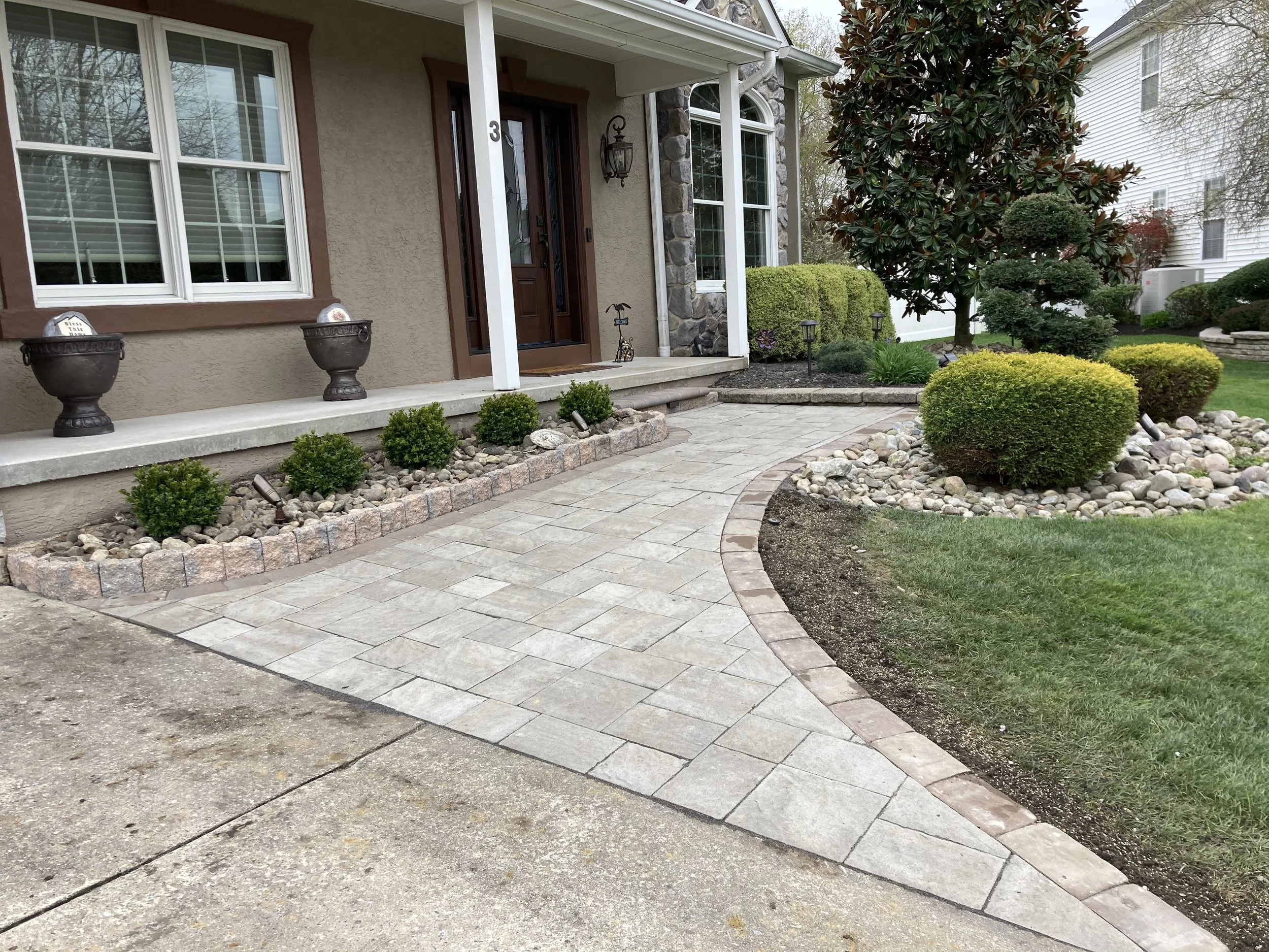 Front yard of a house with a paved walkway, small shrubs, mulch beds, large trees, and neighboring houses in the background.