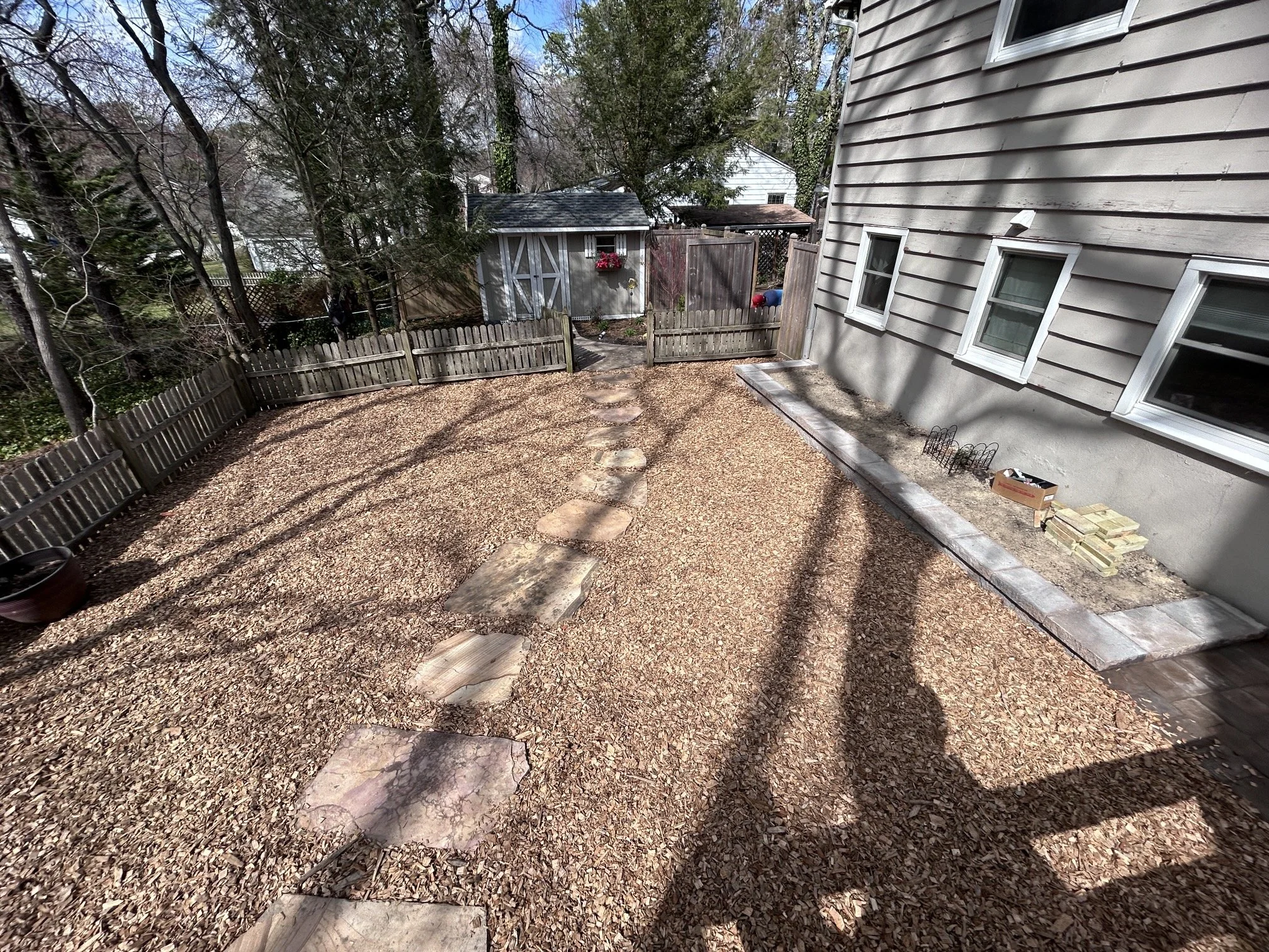 Backyard with mulch ground, stepping stone path, wooden fence, shed with flower basket, trees, house exterior with three small windows, pile of bricks, and garden bed edging.