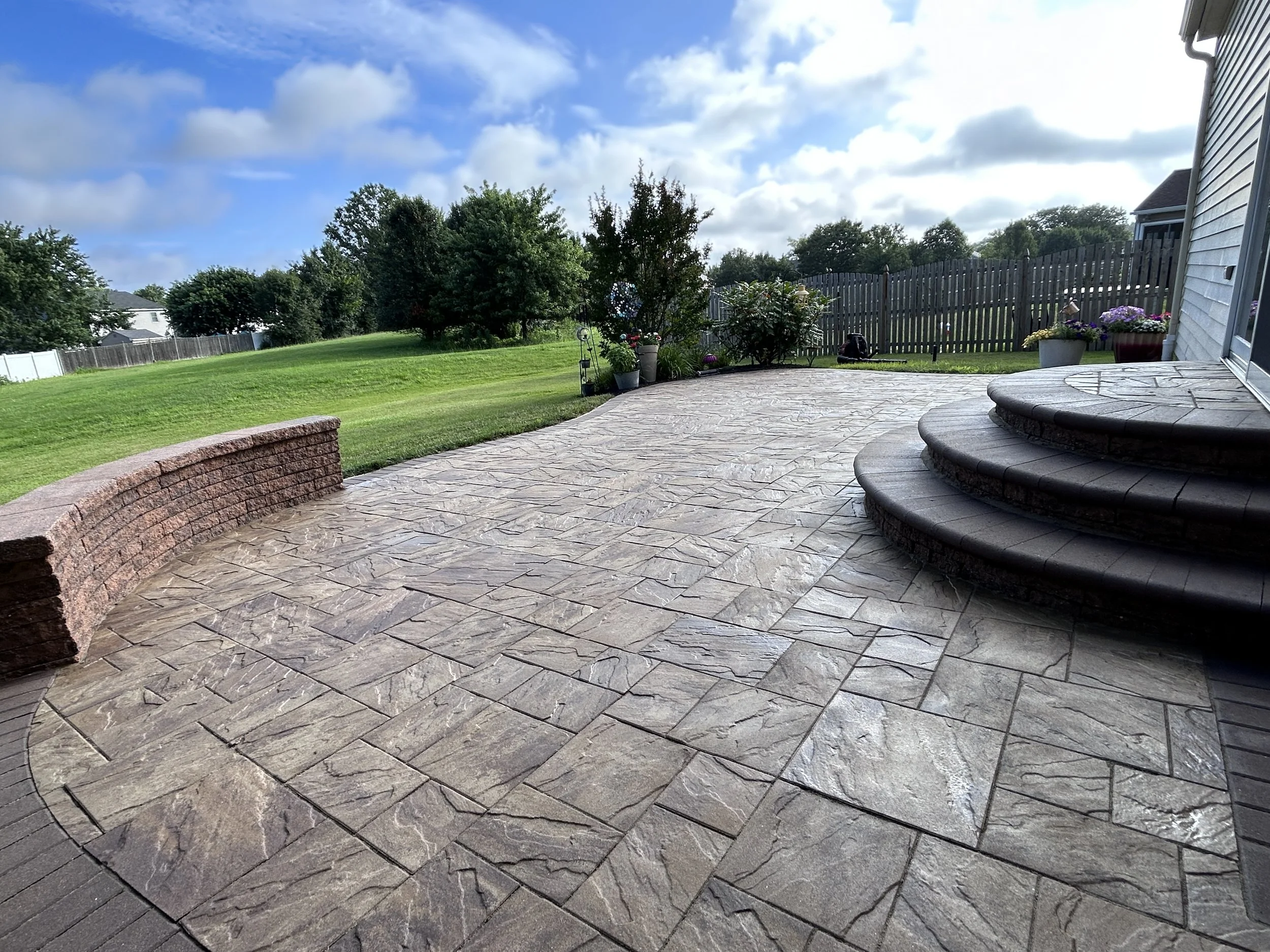 Backyard patio with textured stone flooring, curved brick wall, wooden steps, potted plants, and a grassy yard with trees in the background under a partly cloudy sky.