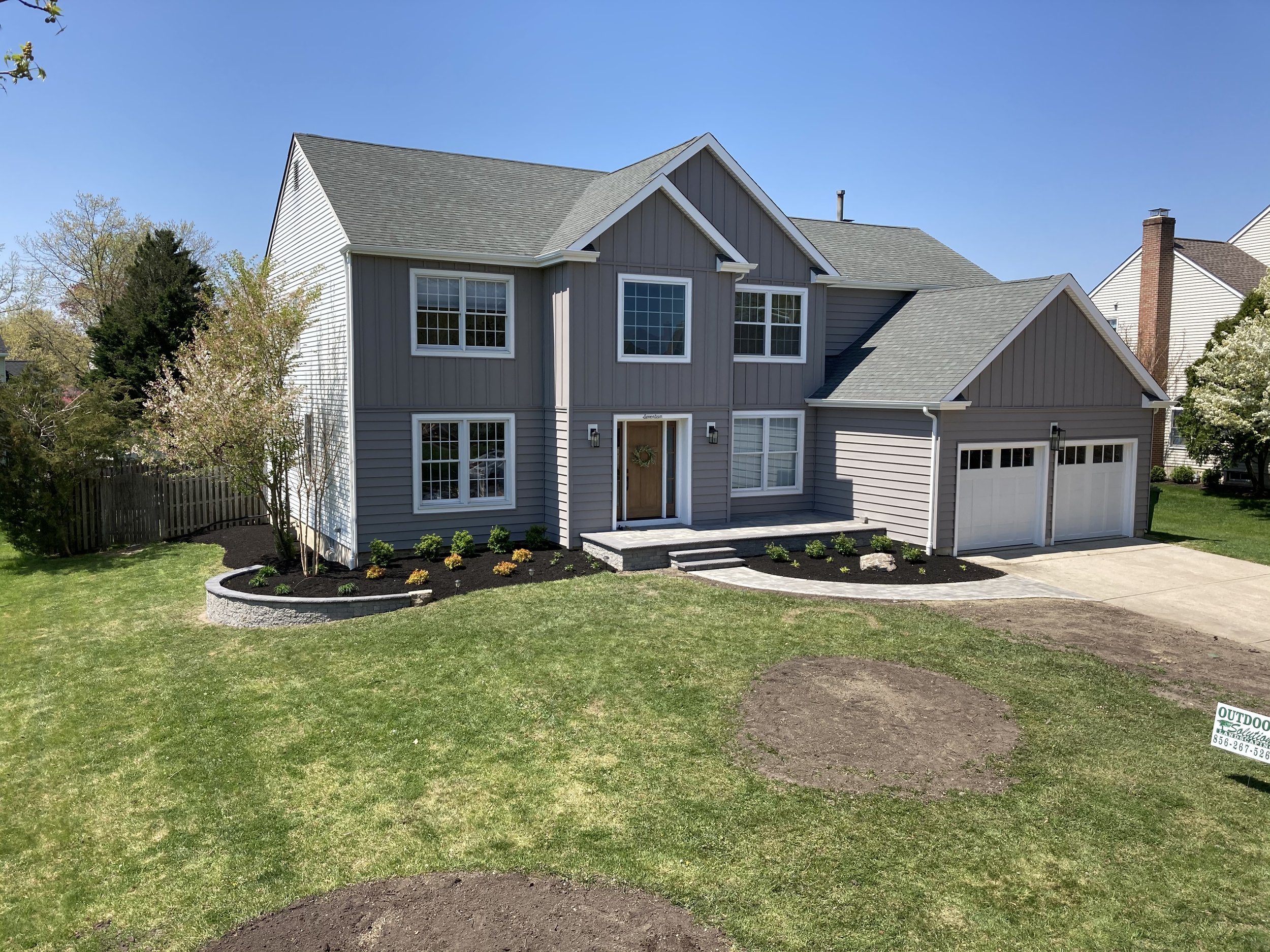 Front view of a two-story house with gray siding, a gable roof, a two-car garage, and a landscaped front lawn with newly planted small bushes and trees.