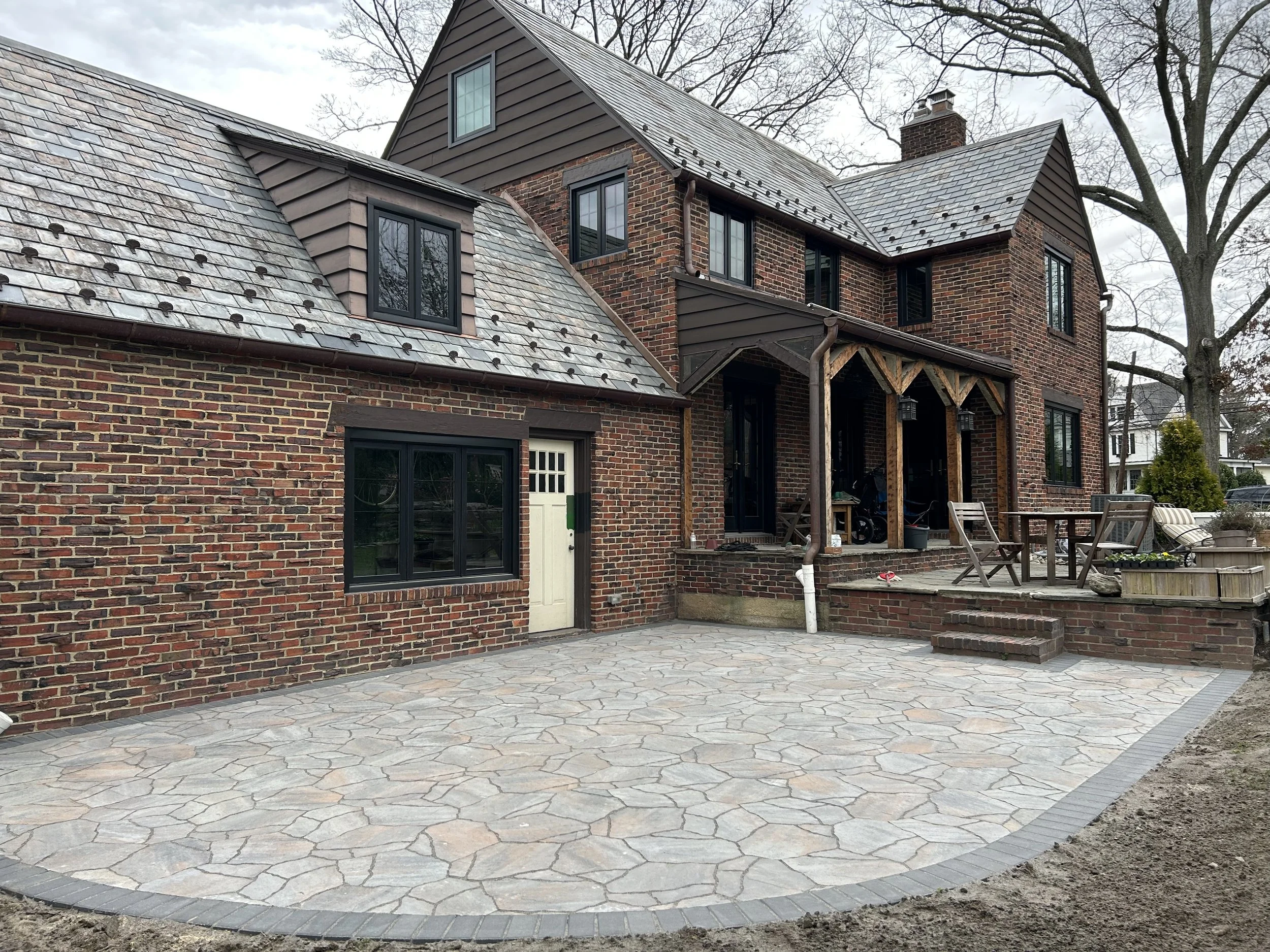 Backyard with a stone patio, brick house, and a large tree in the background.