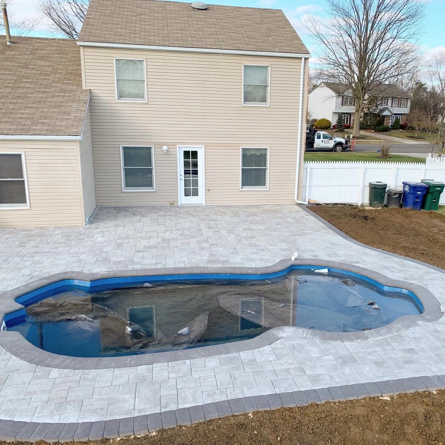 Backyard with an in-ground kidney-shaped swimming pool with a blue liner, surrounded by stone pavers, with some rocks and water in the pool, a beige house with three windows and a door, a white fence, and construction materials.