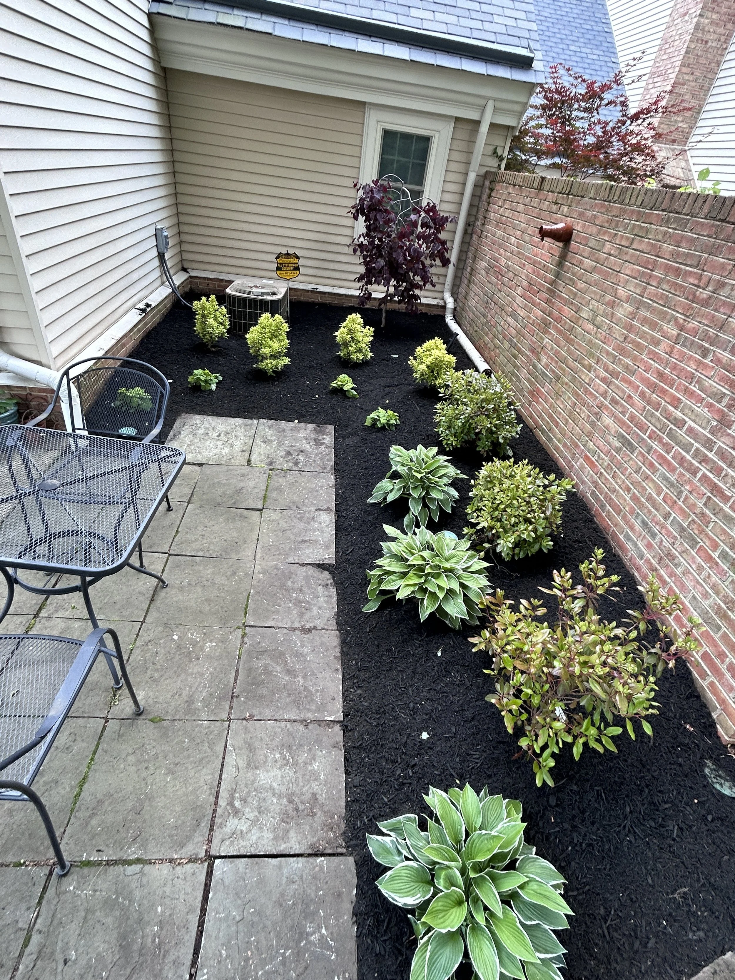 Backyard patio with paved area and garden bed with various plants along a brick wall and house wall.