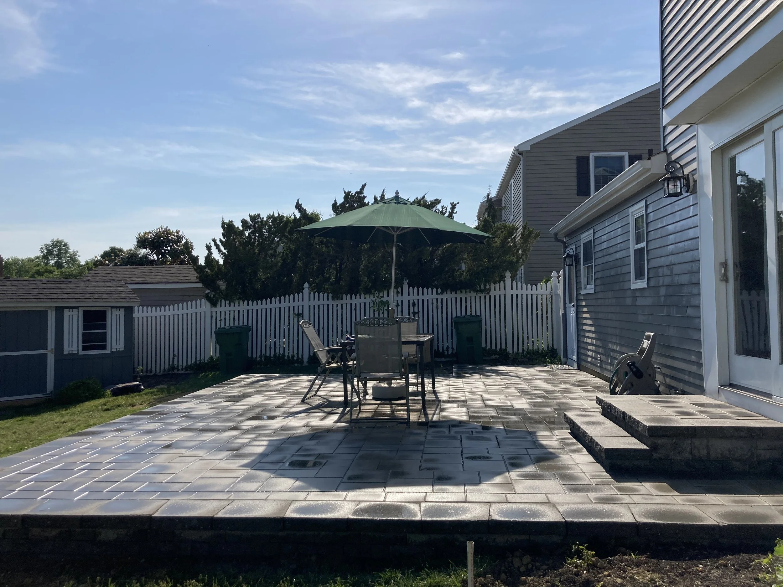 Backyard patio with a green umbrella, table with chairs, two trash bins, a hose reel, a white picket fence, neighboring houses, and a partly cloudy sky.