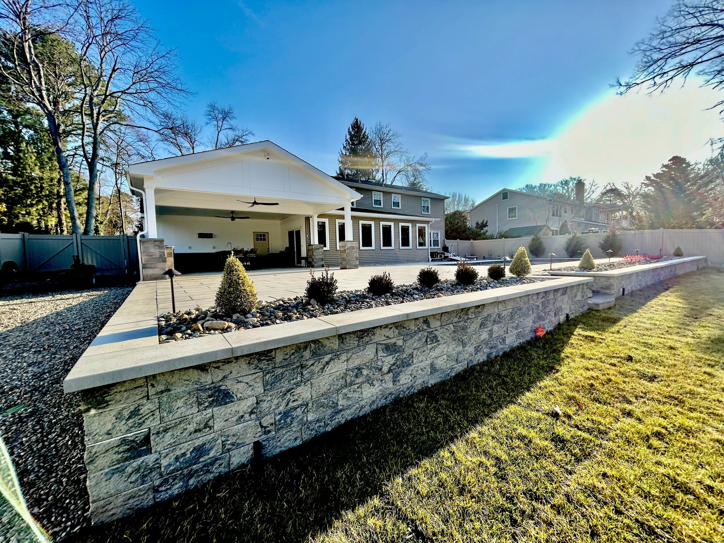 Backyard with a raised stone patio, landscaped with small bushes and decorative rocks, adjacent to a grassy lawn, with a house featuring a covered patio and neighboring houses under a bright, sunny sky.
