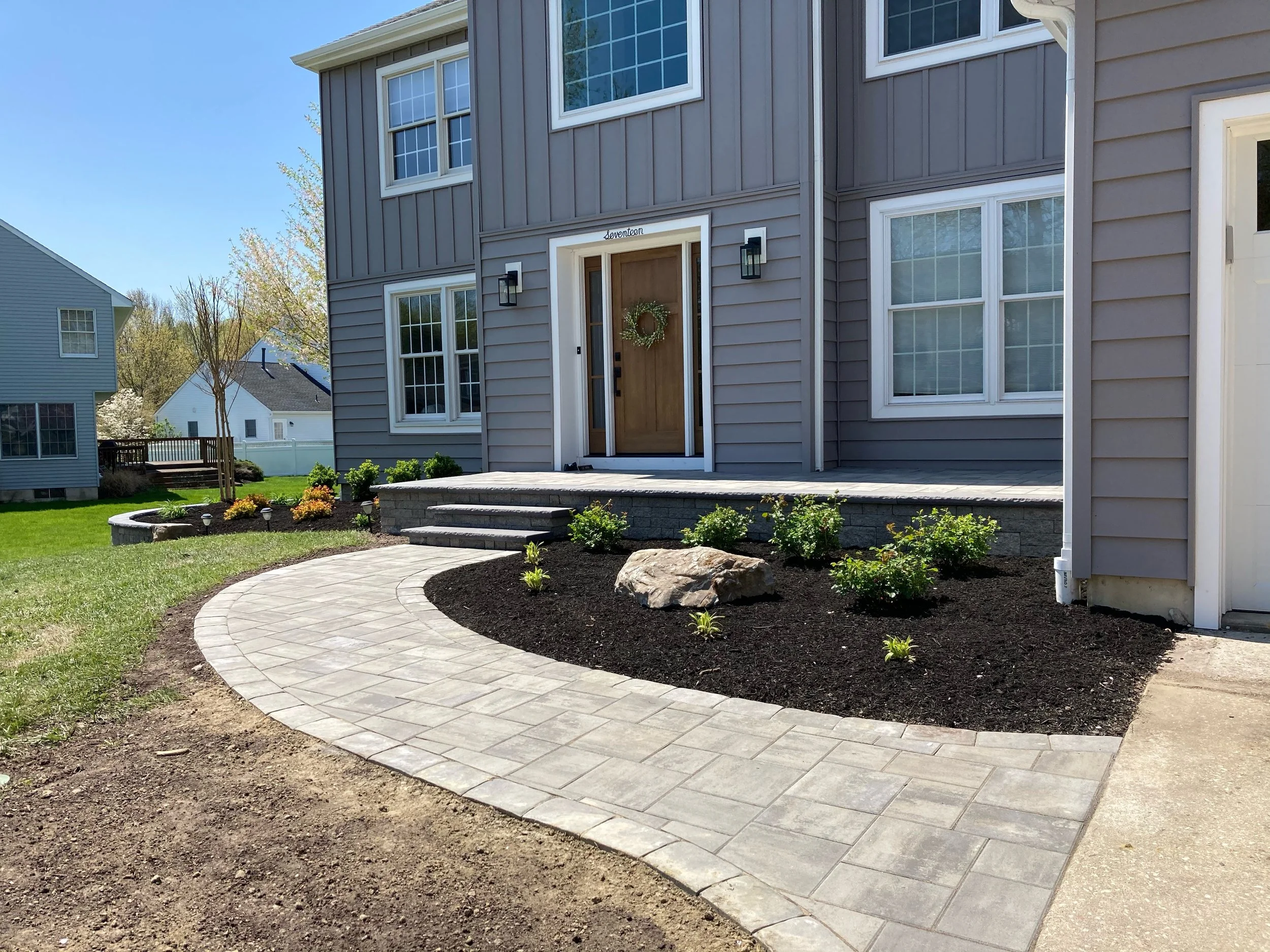 Newly landscaped backyard with a curved concrete paver patio, small plants, and a house with a wooden front door decorated with a wreath.
