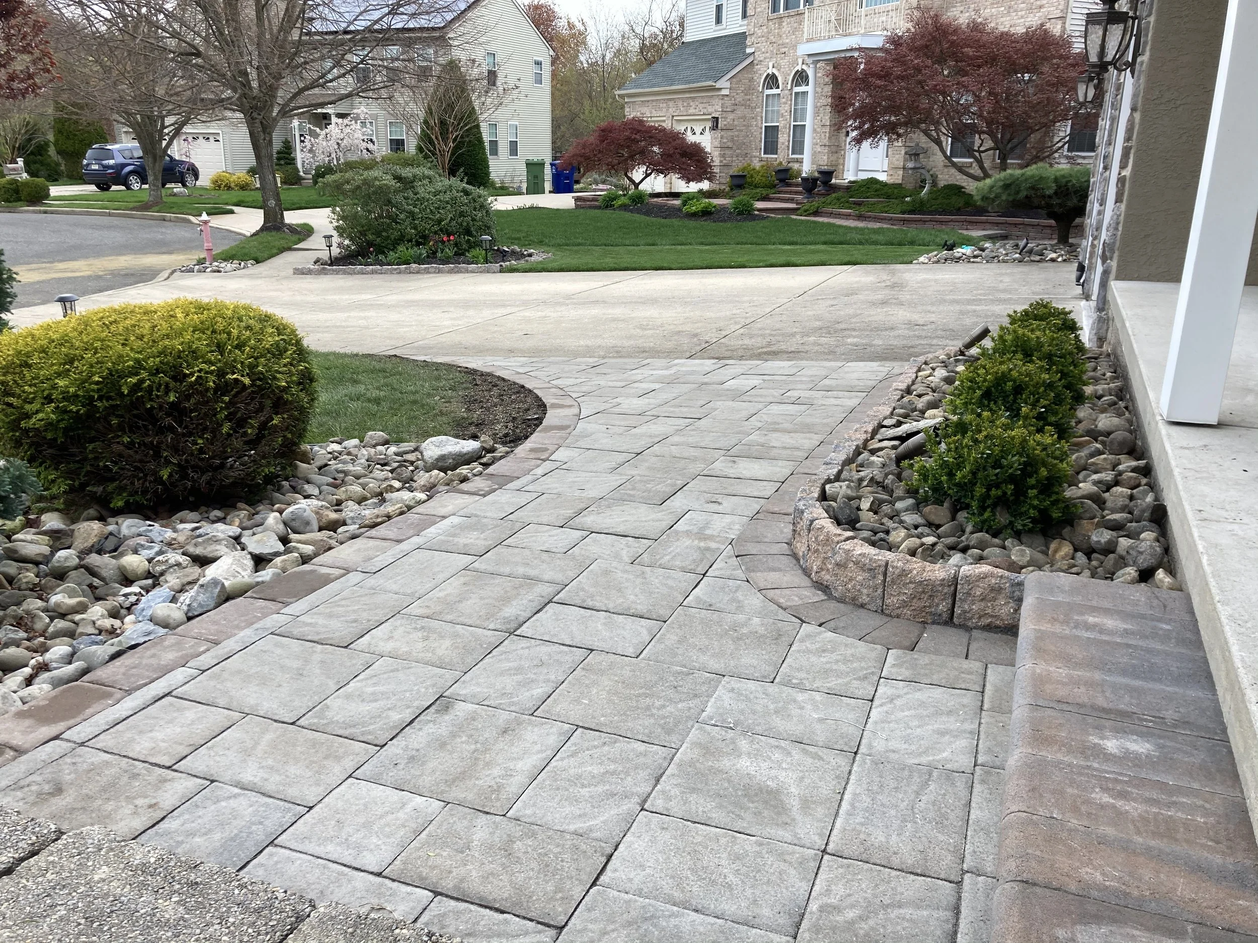 View of a residential front yard with a concrete driveway, landscaped garden beds with rocks and shrubs, and houses in the background.