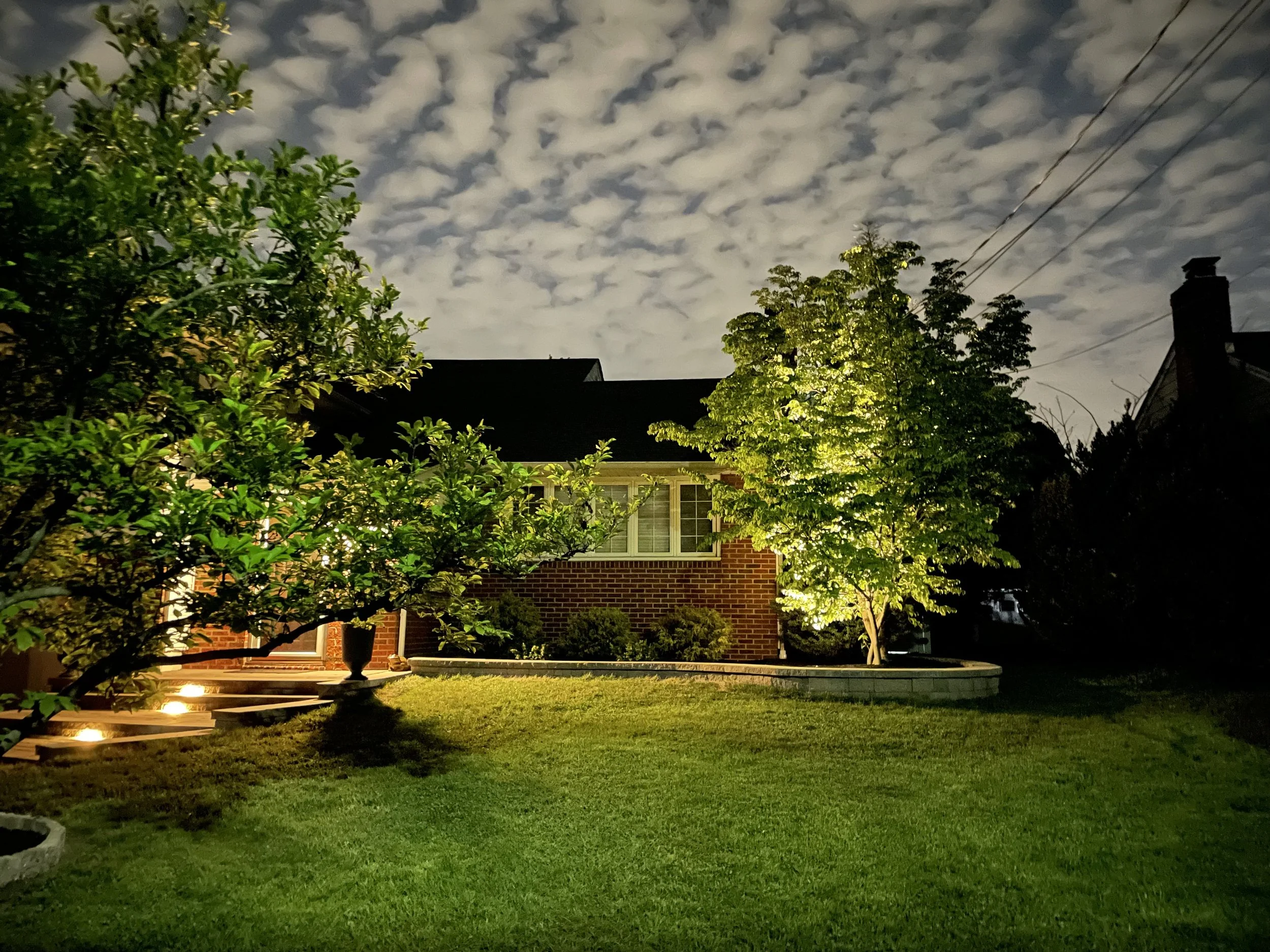 Nighttime view of a suburban backyard with a lawn, trees illuminated by outdoor lights, a brick house with windows, and a cloudy sky overhead.