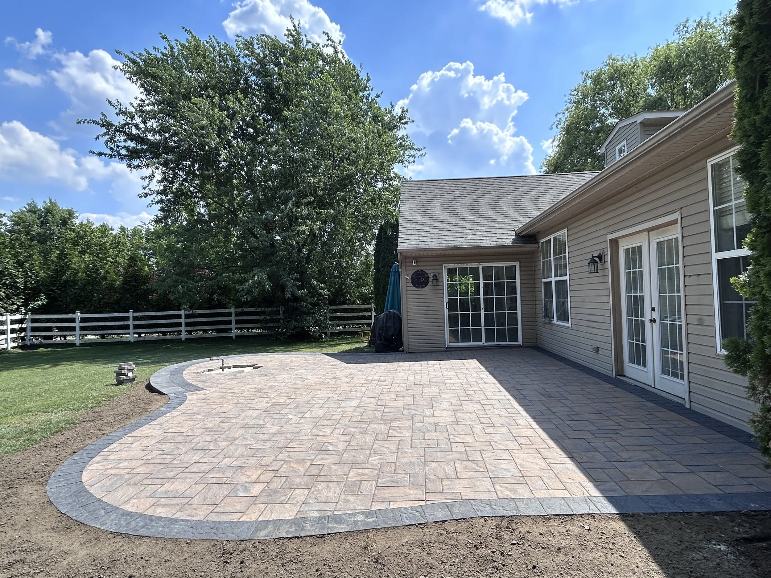 Backyard patio with stamped concrete, adjacent to a beige house with sliding glass doors and windows, surrounded by green trees under a partly cloudy sky.