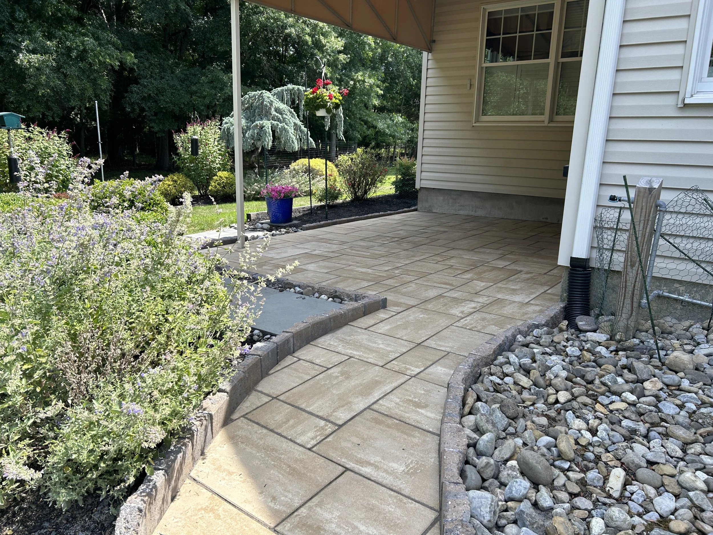 A backyard patio with a beige tiled floor, bordered by curved stone edging. To the left, there are flowering plants with purple and white blooms. In the background, there is a lush green garden with trees and bushes. A hanging flower basket is suspen