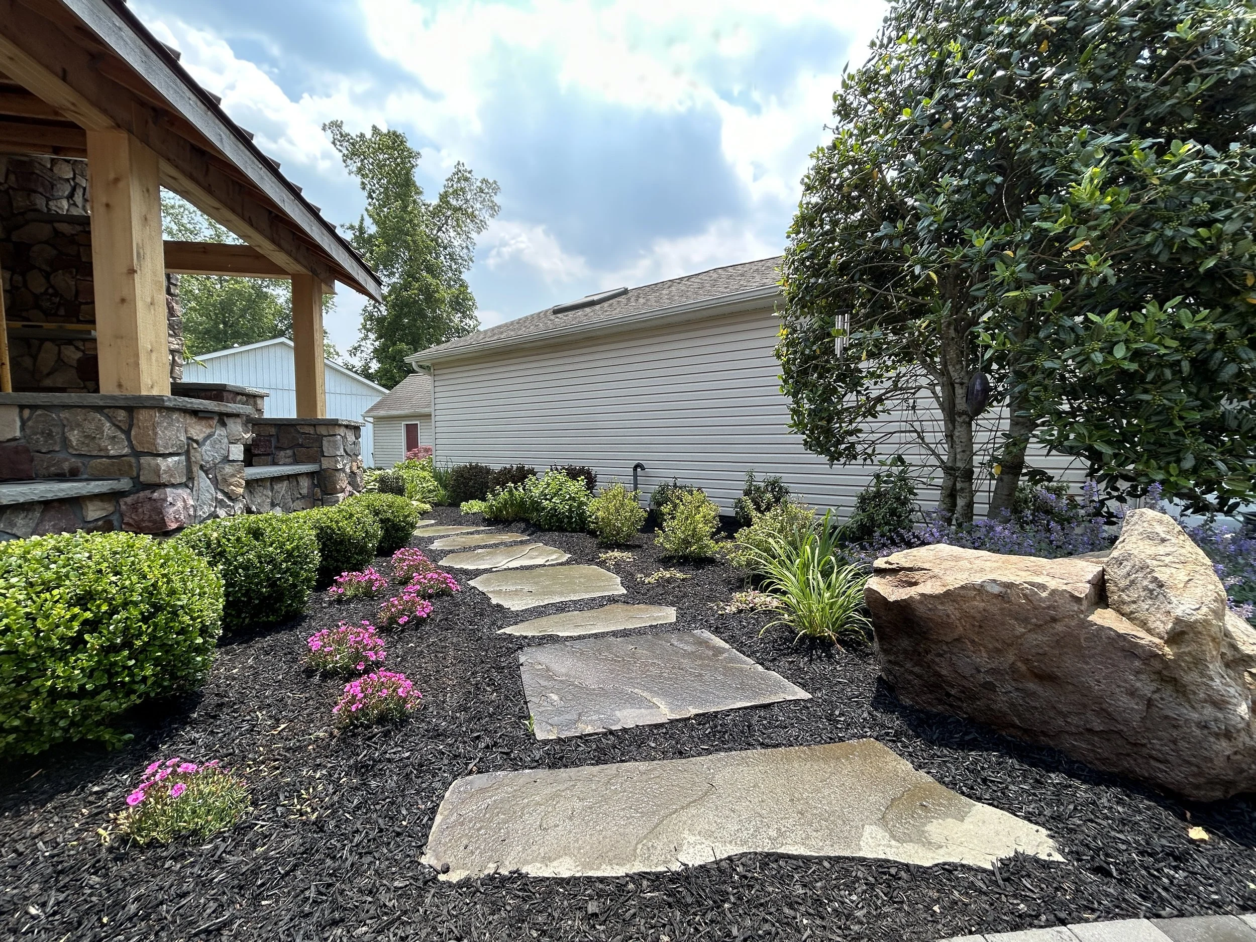 A landscaped backyard with a stone pathway, bushes, flowering plants, a large rock, and a tree, with a white house and a wooden patio structure nearby.