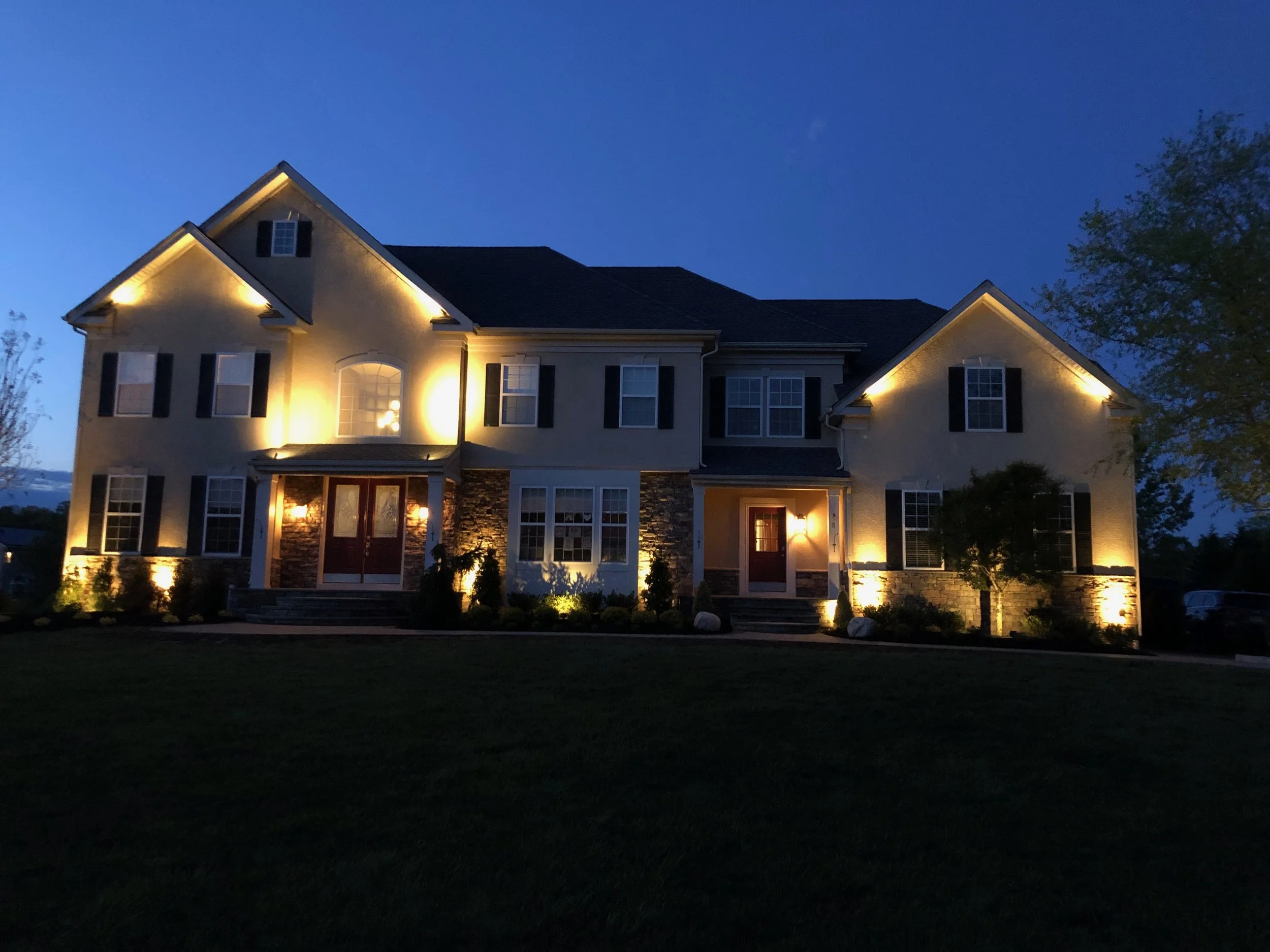 A large, two-story house illuminated with exterior lighting at dusk, with a manicured front yard and trees nearby.