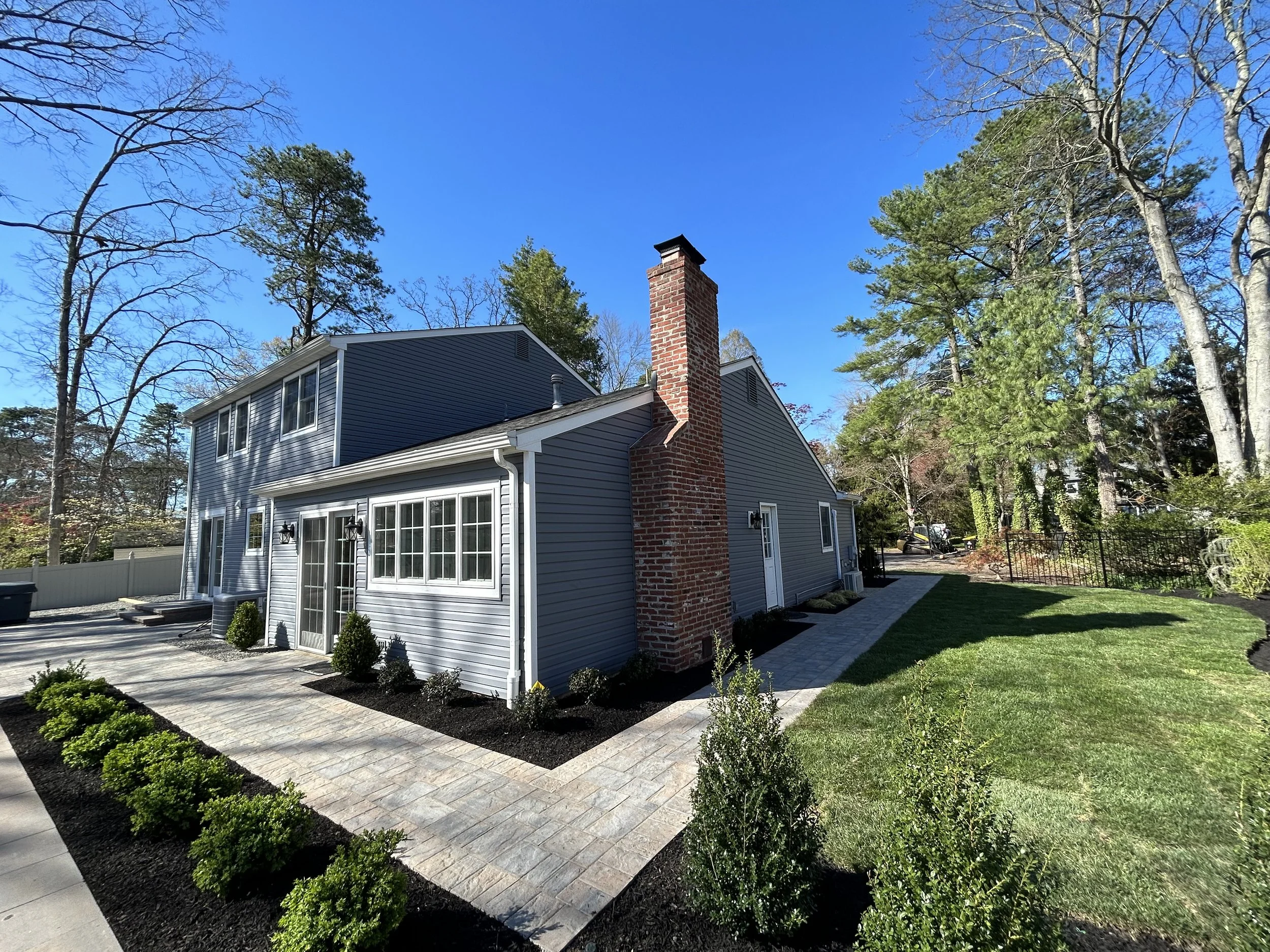 Backyard of a house with a brick chimney, gray siding, large windowed doors, well-maintained lawn, paved walkways, and trees in the background on a sunny day.