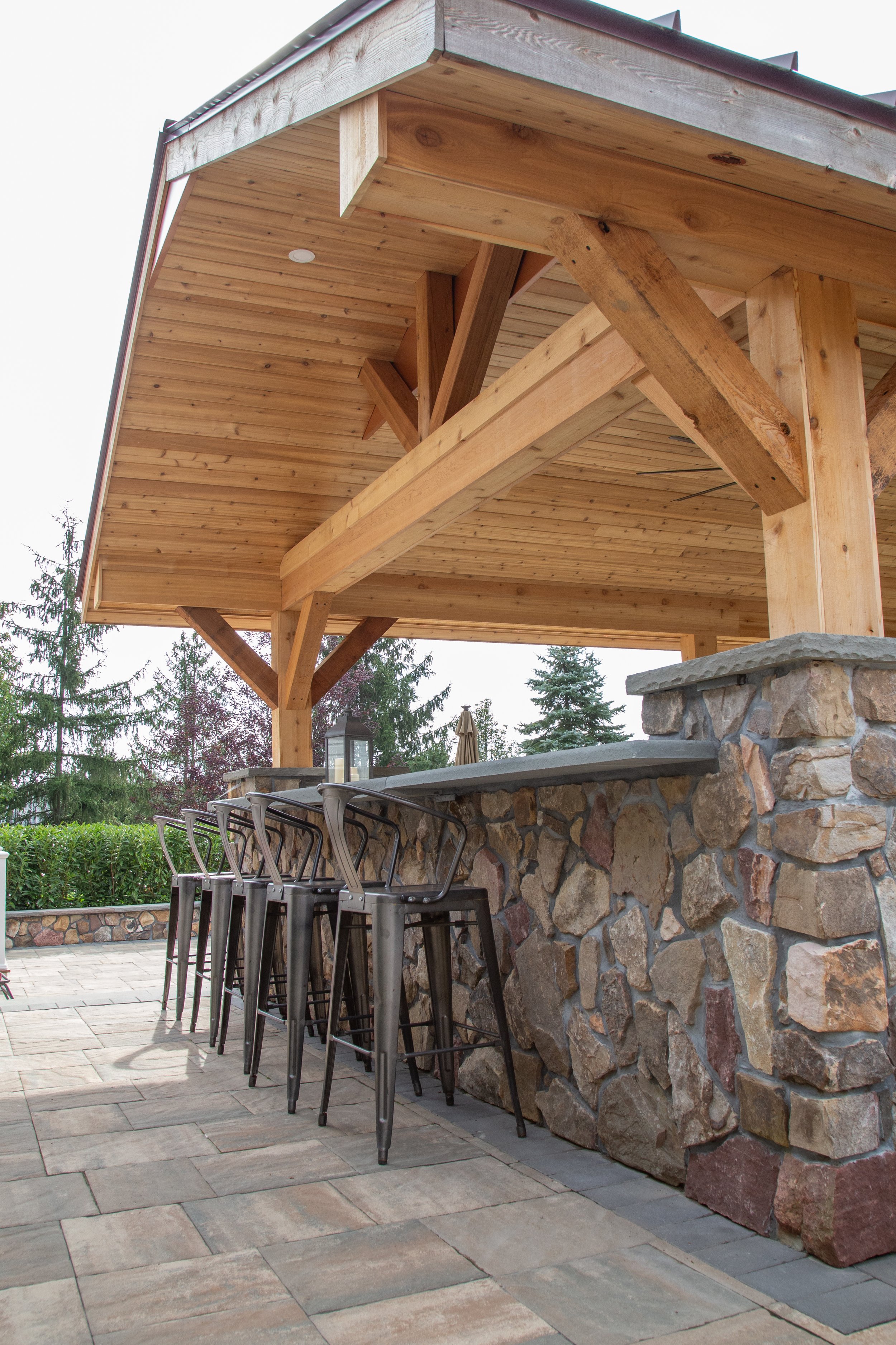 Outdoor bar area with a stone counter, wooden ceiling and support beams, and four black metal barstools, set on a paved patio with trees in the background.