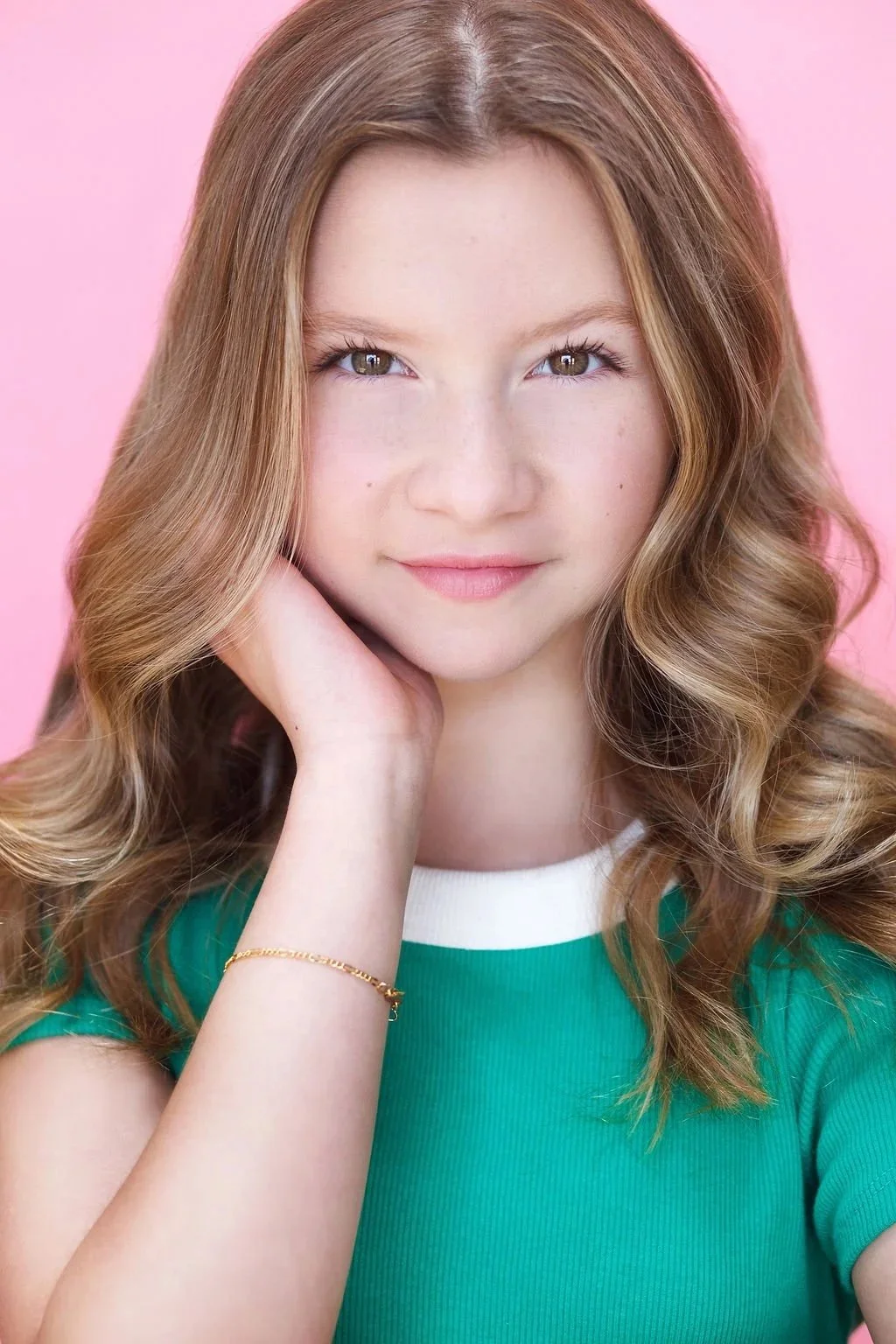Portrait of a young girl with long wavy brown hair, green eyes, and light skin, posing with her hand resting on her cheek against a pink background.