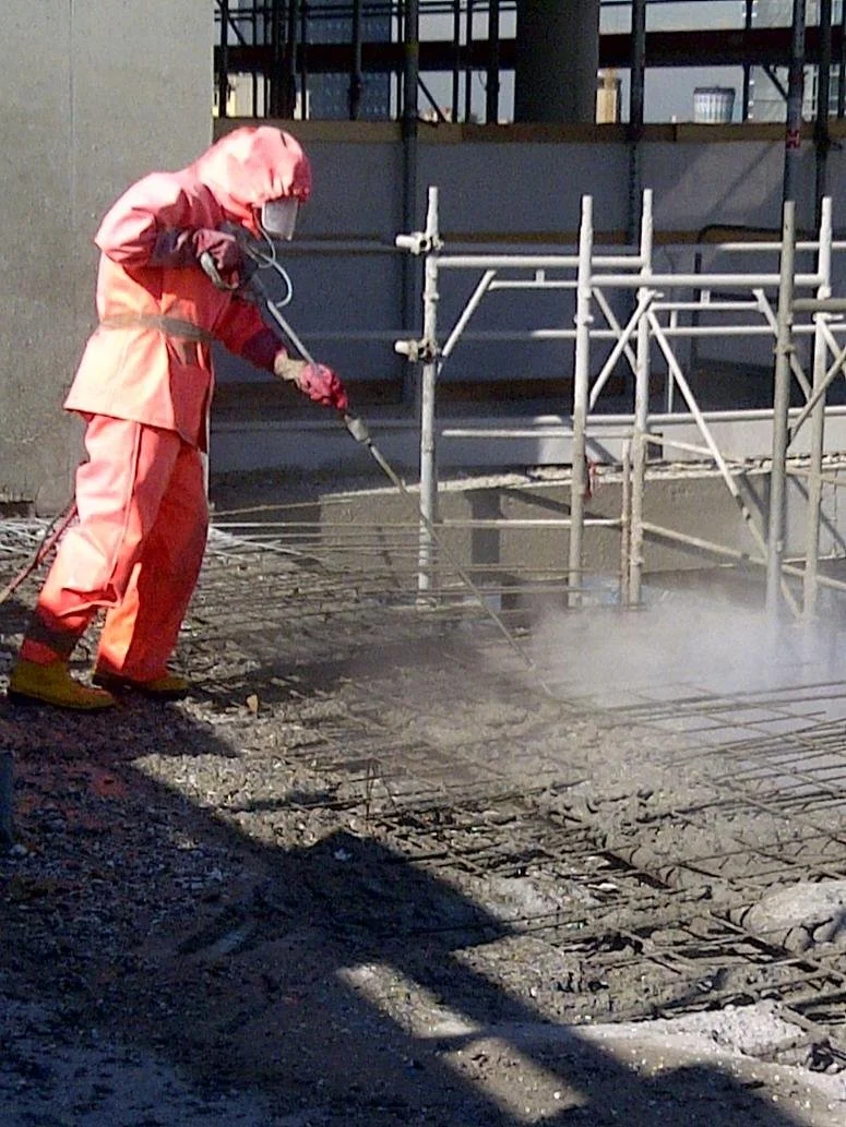Una persona in tuta protettiva arancione utilizza un'idropulitrice su una superficie cementata in un cantiere, vicino a impalcature.
