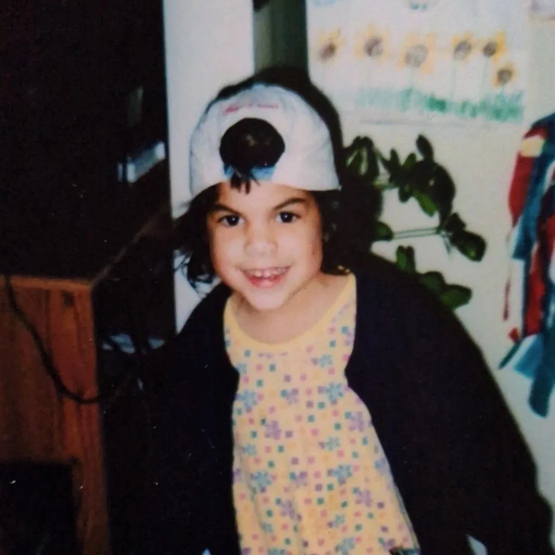 A young girl smiling, wearing a baseball cap backwards and a colorful dress, standing in a room with a plant and a paper with writing and images on the wall behind her.