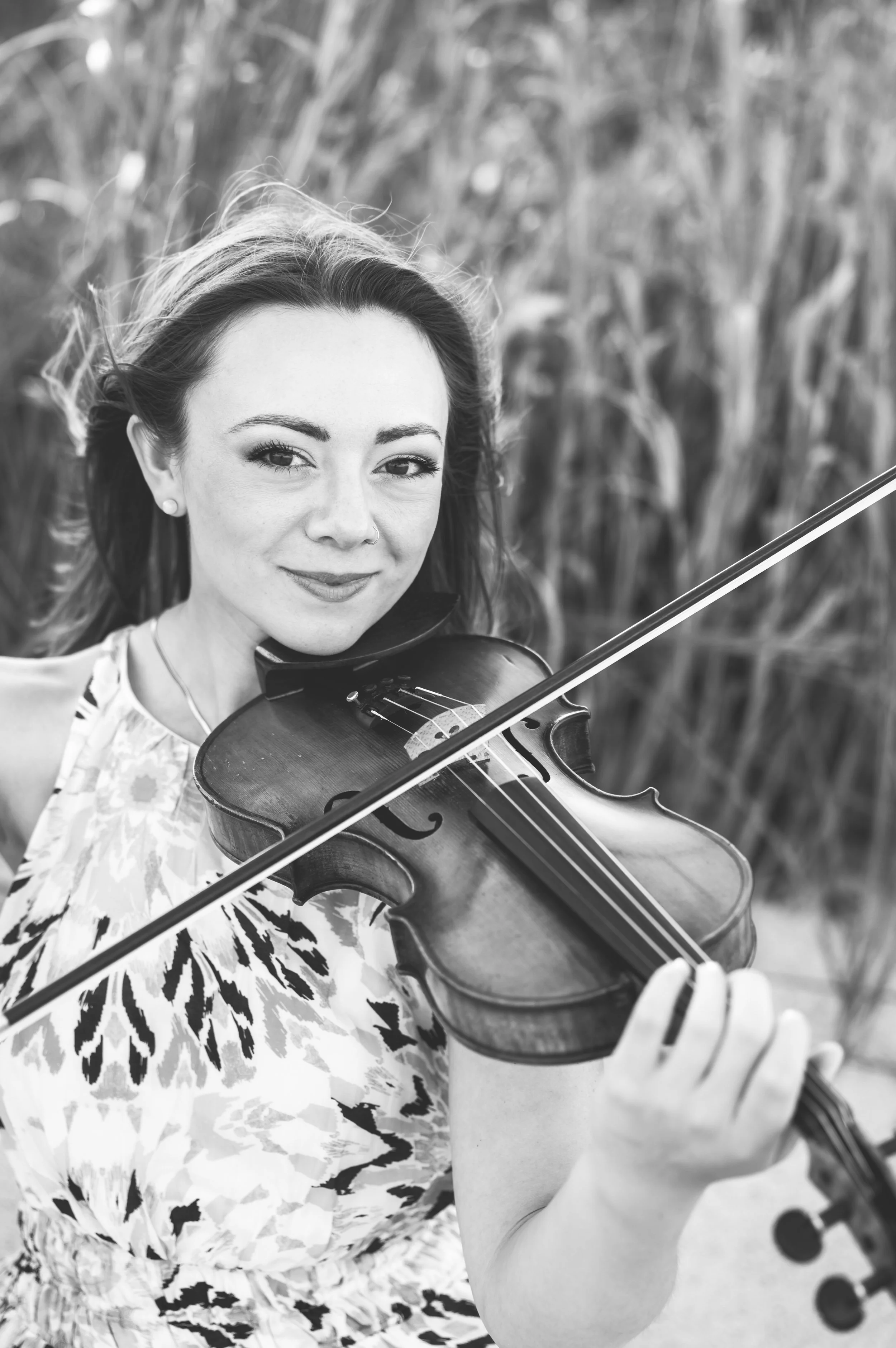 A woman with long hair playing a violin outdoors, smiling, with a blurred background of tall grass.