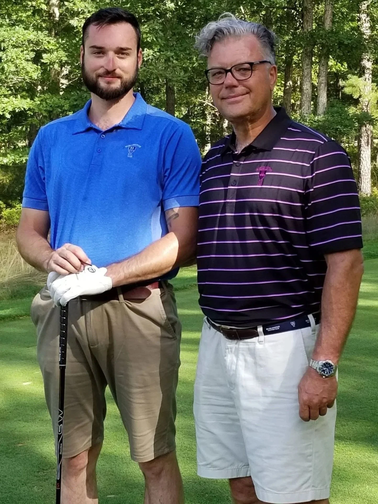 Two men on a golf course, standing side by side outdoors with trees in the background.
