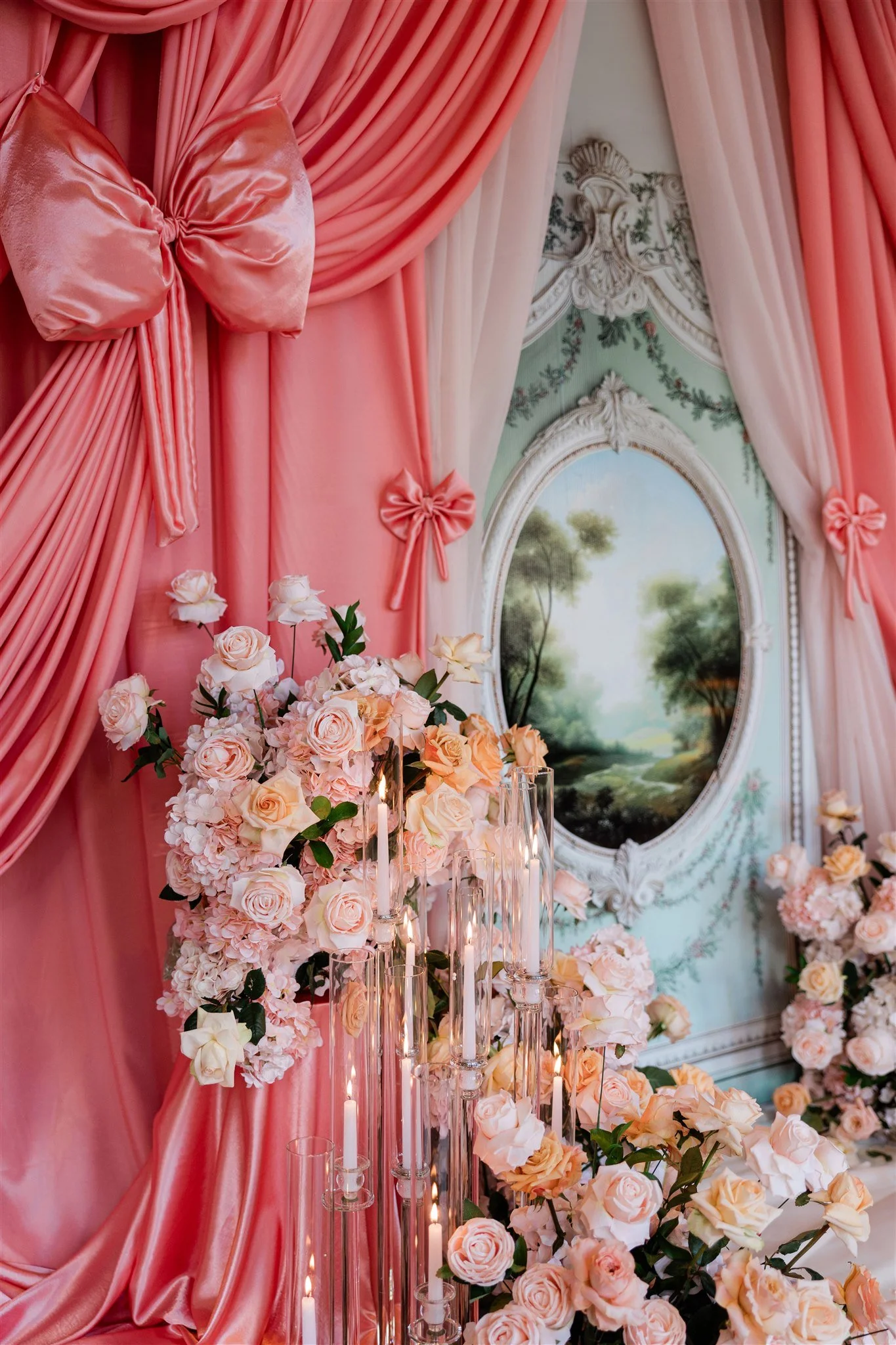 Elegant pink drapery with bows, floral arrangements with pink roses, and tall glass candle holders with lit candles, set against a decorative wall with a landscape painting.
