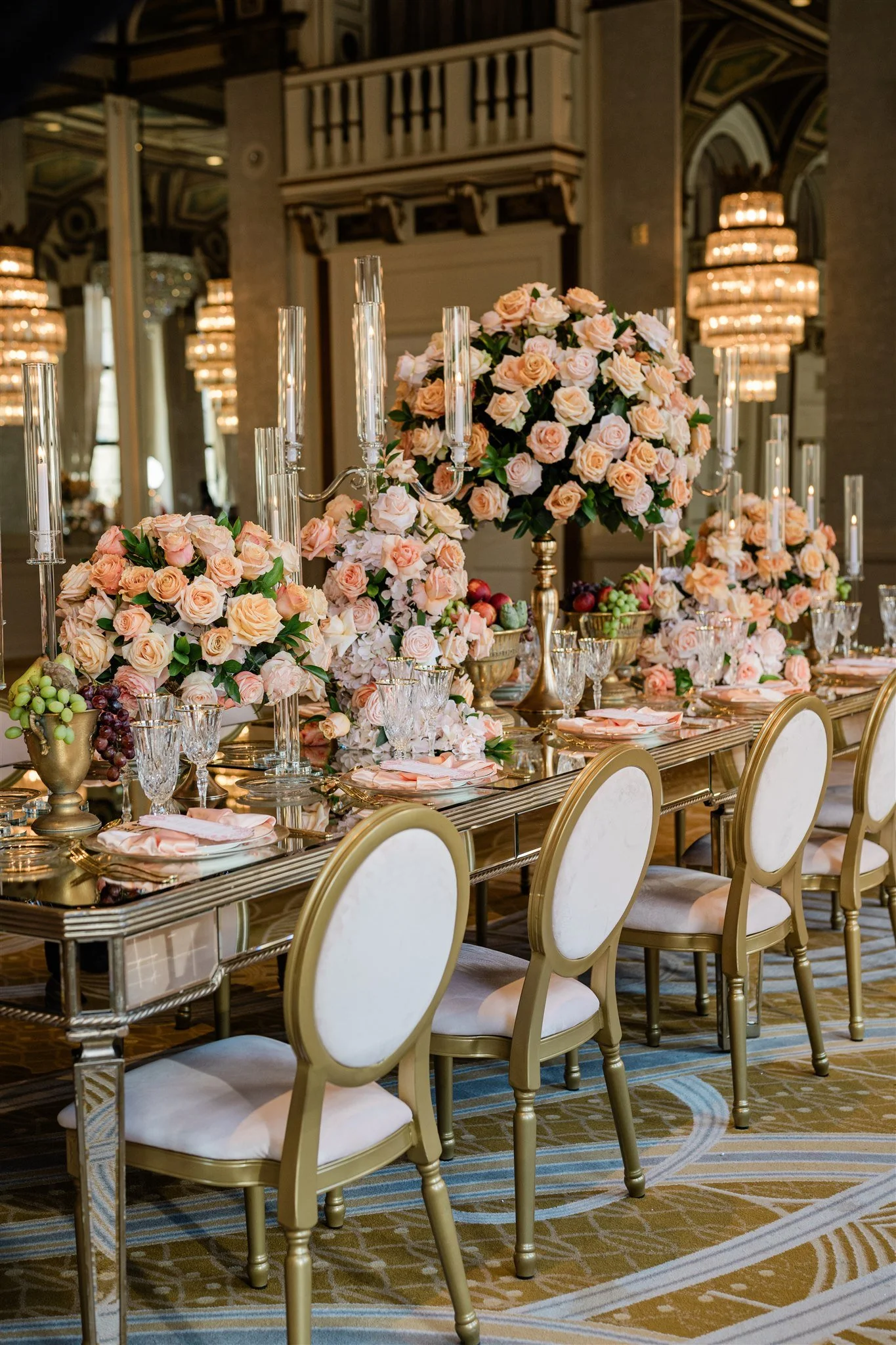 Elegant banquet table decorated with large floral arrangements of pink and peach roses, surrounded by gold and white chairs, crystal glassware, and tall candelabras, in a luxurious room with chandeliers and ornate architectural details.