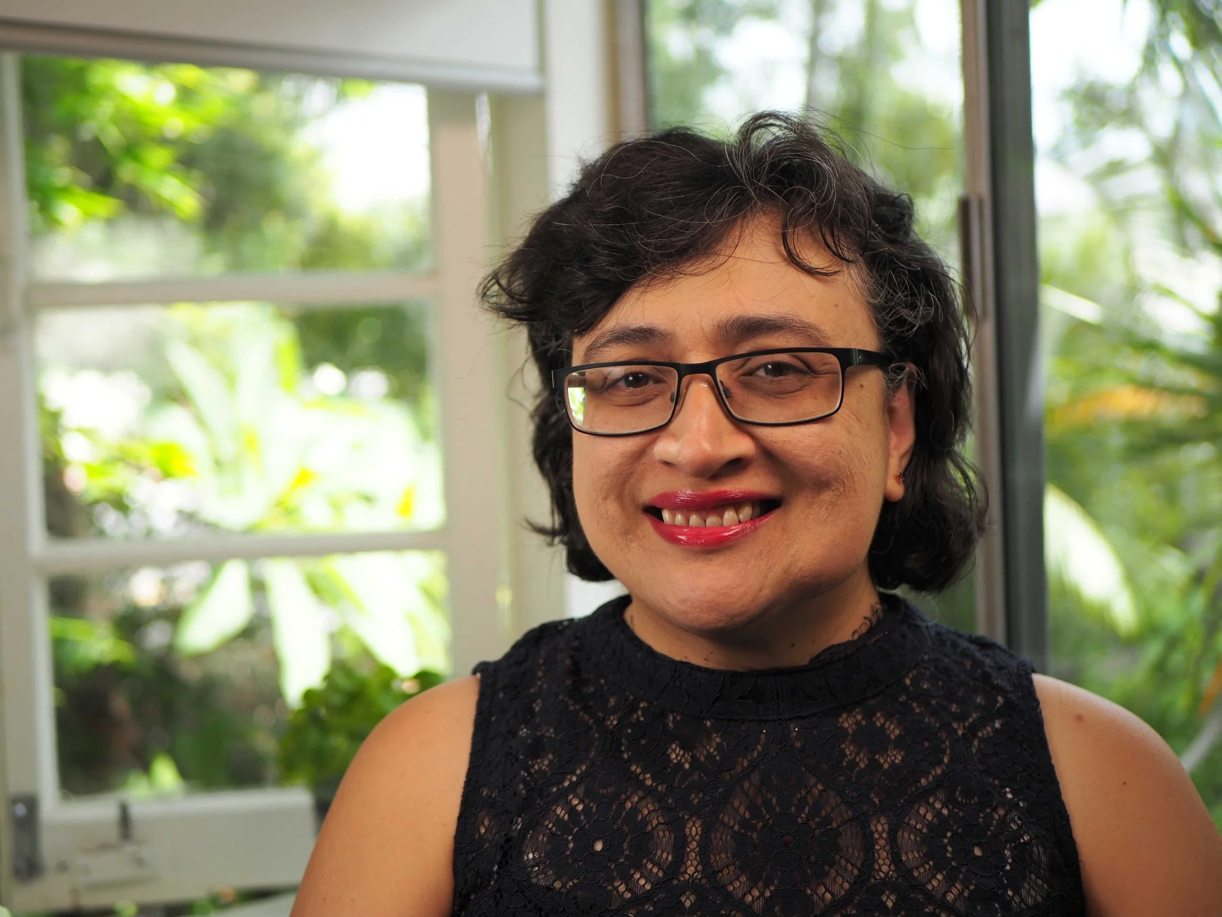 A woman with short curly dark hair, wearing glasses and a sleeveless lacy black top, smiling indoors in front of a window with green foliage outside.
