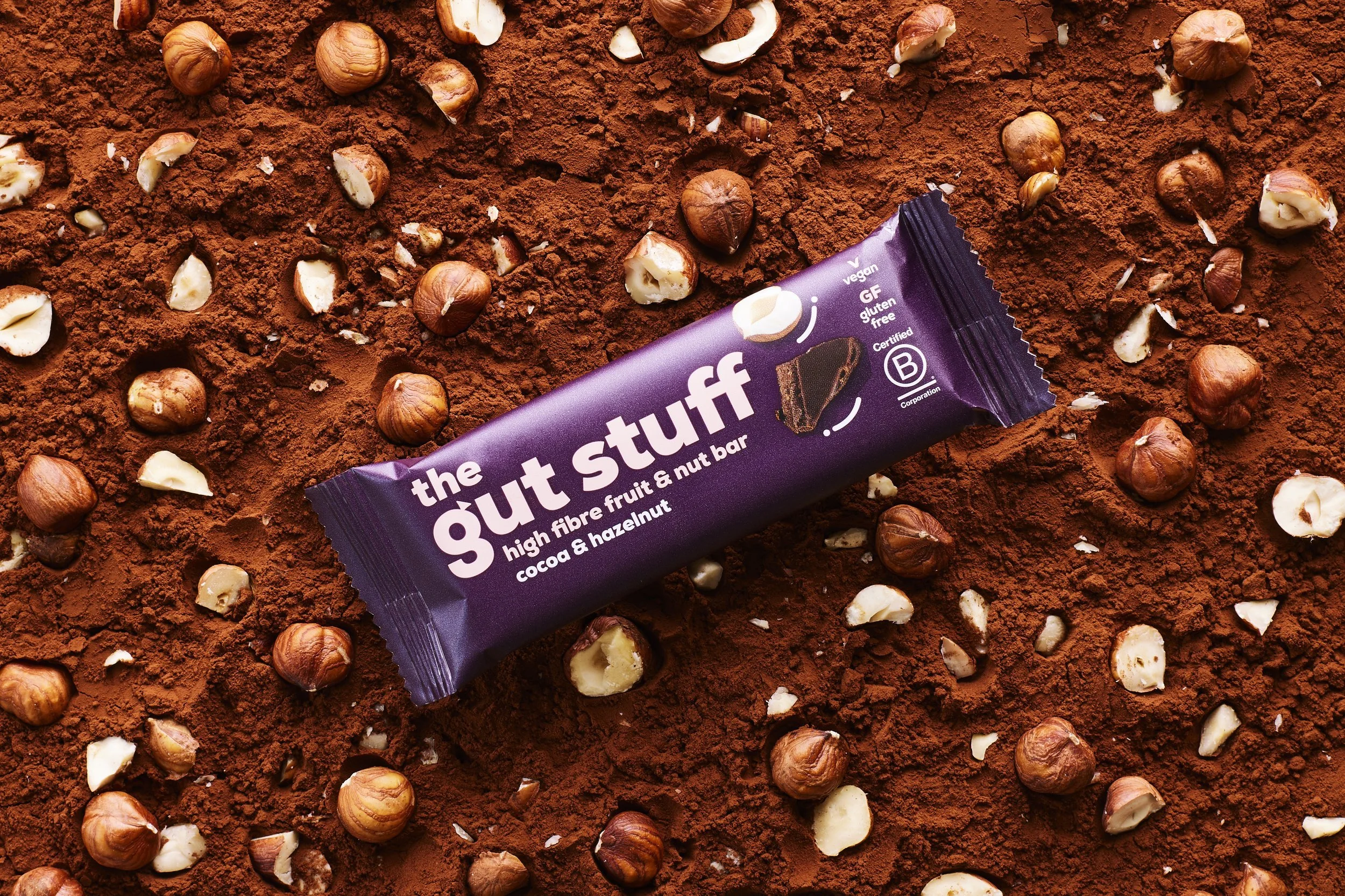 A bar of dark chocolate-flavored snack called 'the guilt stuff' surrounded by scattered hazelnuts on a bed of cocoa powder.