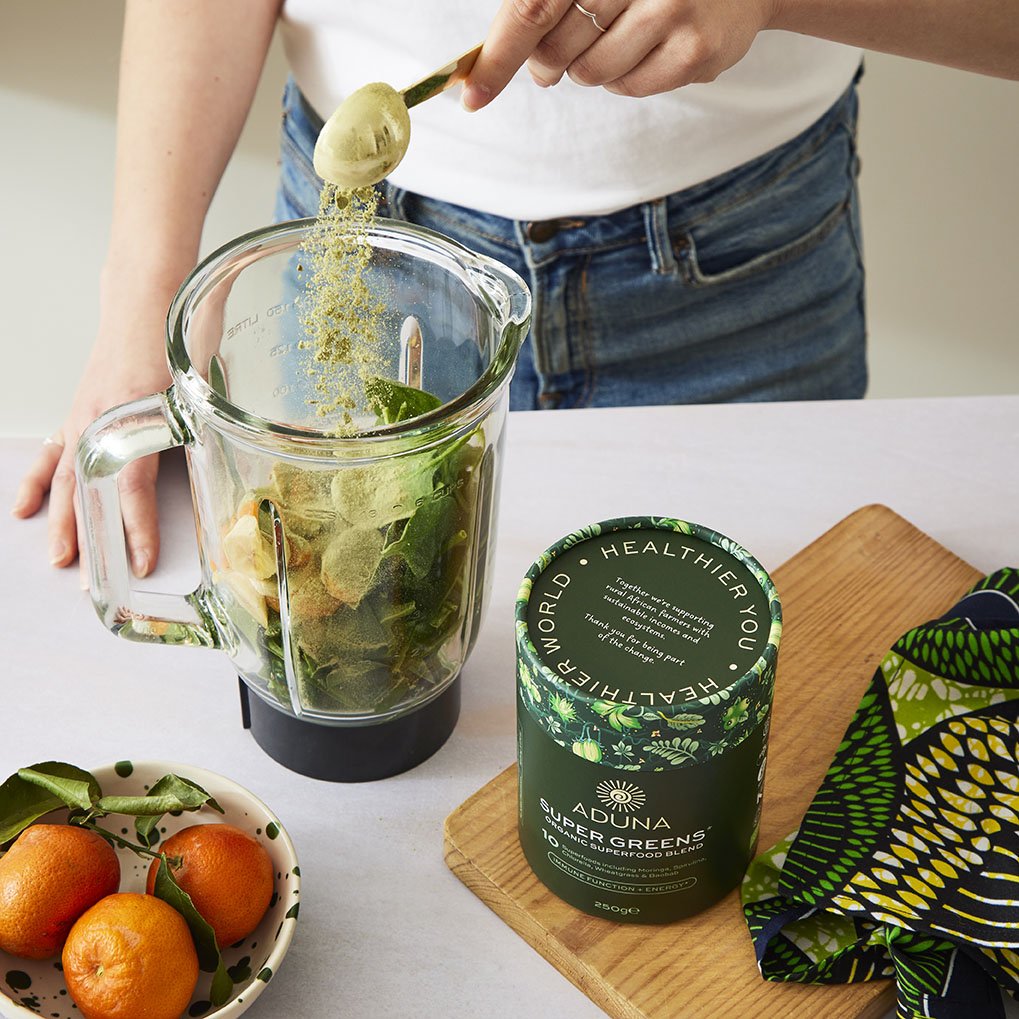 Person preparing a green smoothie with fresh fruits and leafy greens in a blender. A bowl of oranges is on the table, and a container of super greens powder is nearby.