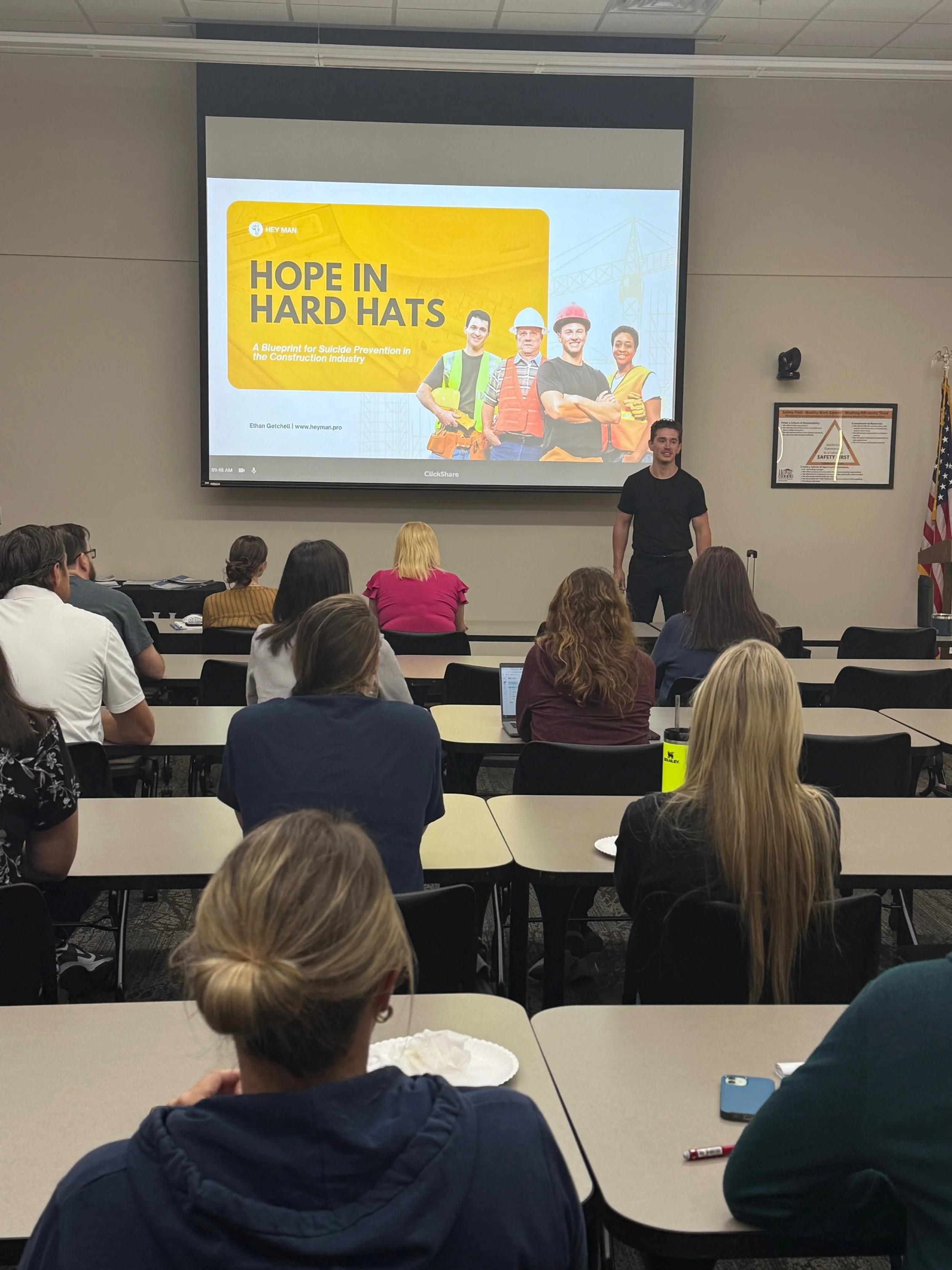 A classroom presentation with a projector screen displaying a slide titled "Hope in Hard Hats" and a group of construction workers. The presenter is standing near the screen, and the audience is seated at desks, watching and taking notes.