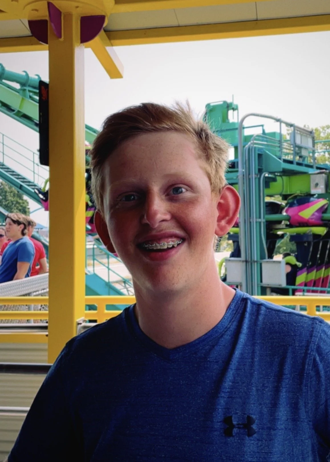A young man with light skin, red hair, braces, and wearing a blue shirt, smiling at an amusement park or carnival. In the background, there are colorful rides and other people.