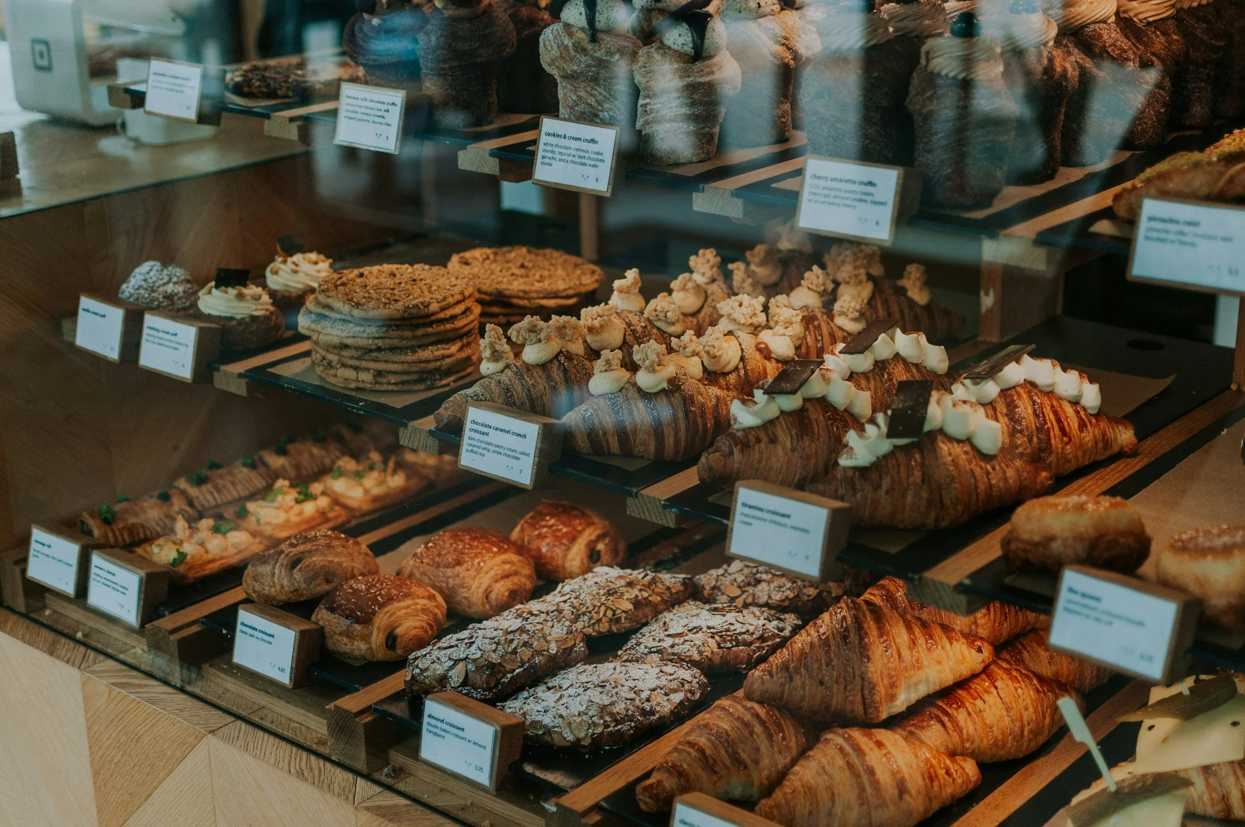 Display case full of various pastries including croissants, cookies, danishes, and muffins in a bakery shop.