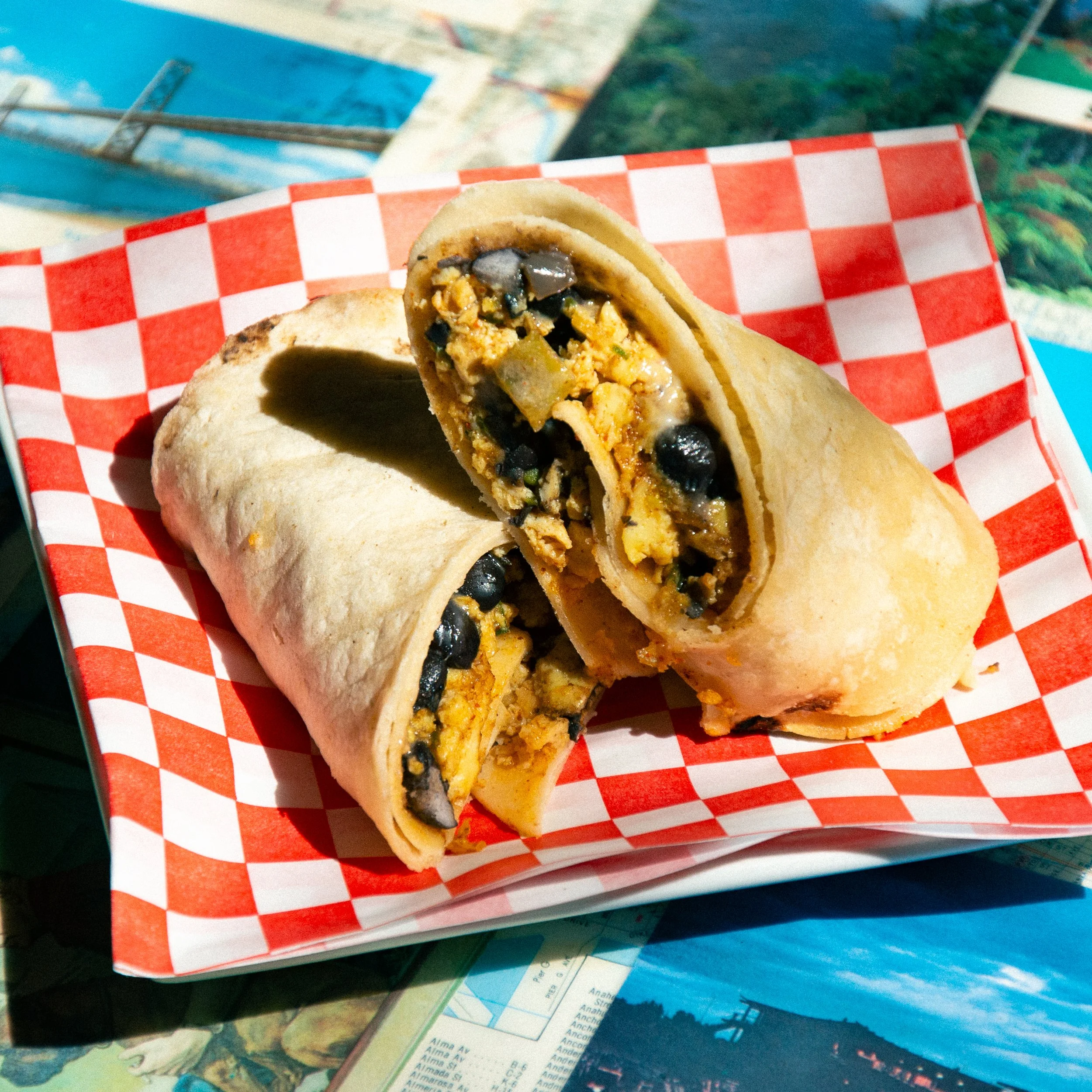 A burrito cut in half, filled with beans, cheese, vegetables, and meat, on a red and white checkered paper inside a basket.