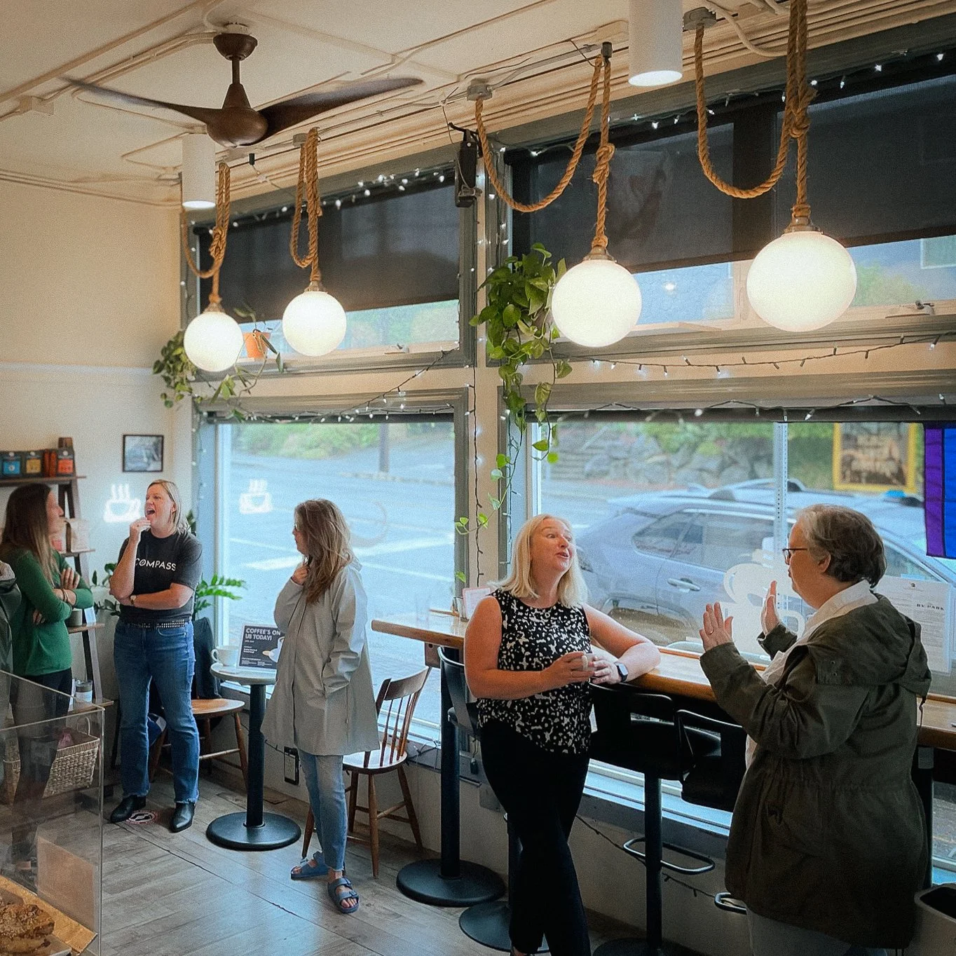 Four women and one man inside a cozy cafe, some sitting and some standing, engaging in conversation, with large windows, hanging globe lights, and green plants.