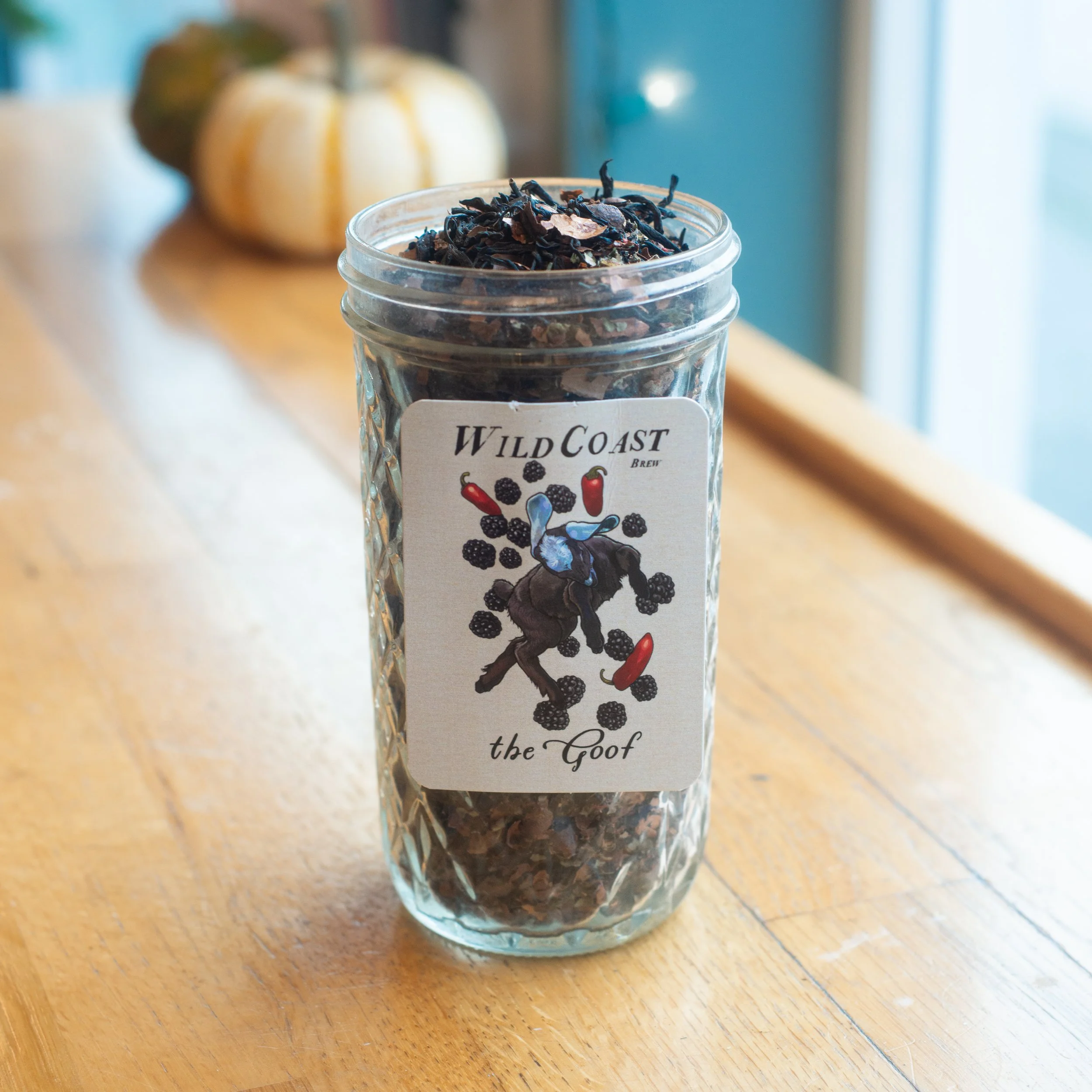 A glass jar filled with dried black tea leaves on a wooden surface, with a label reading 'Wild Coast Brew the Goat' and an illustration of a goat surrounded by blackberries and red peppers, with a pumpkin in the background near a window.