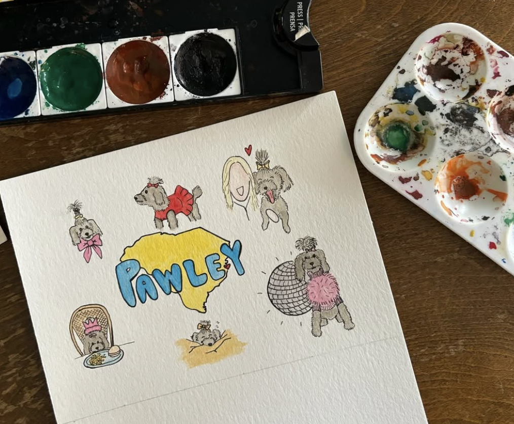 A watercolor illustration of a dog named Pawley surrounded by various outfits and accessories, including a pink dress, a disco ball costume, and a crown, with a map of South Carolina and a woman with blonde hair. There are paint palettes and watercolors beside the drawing.