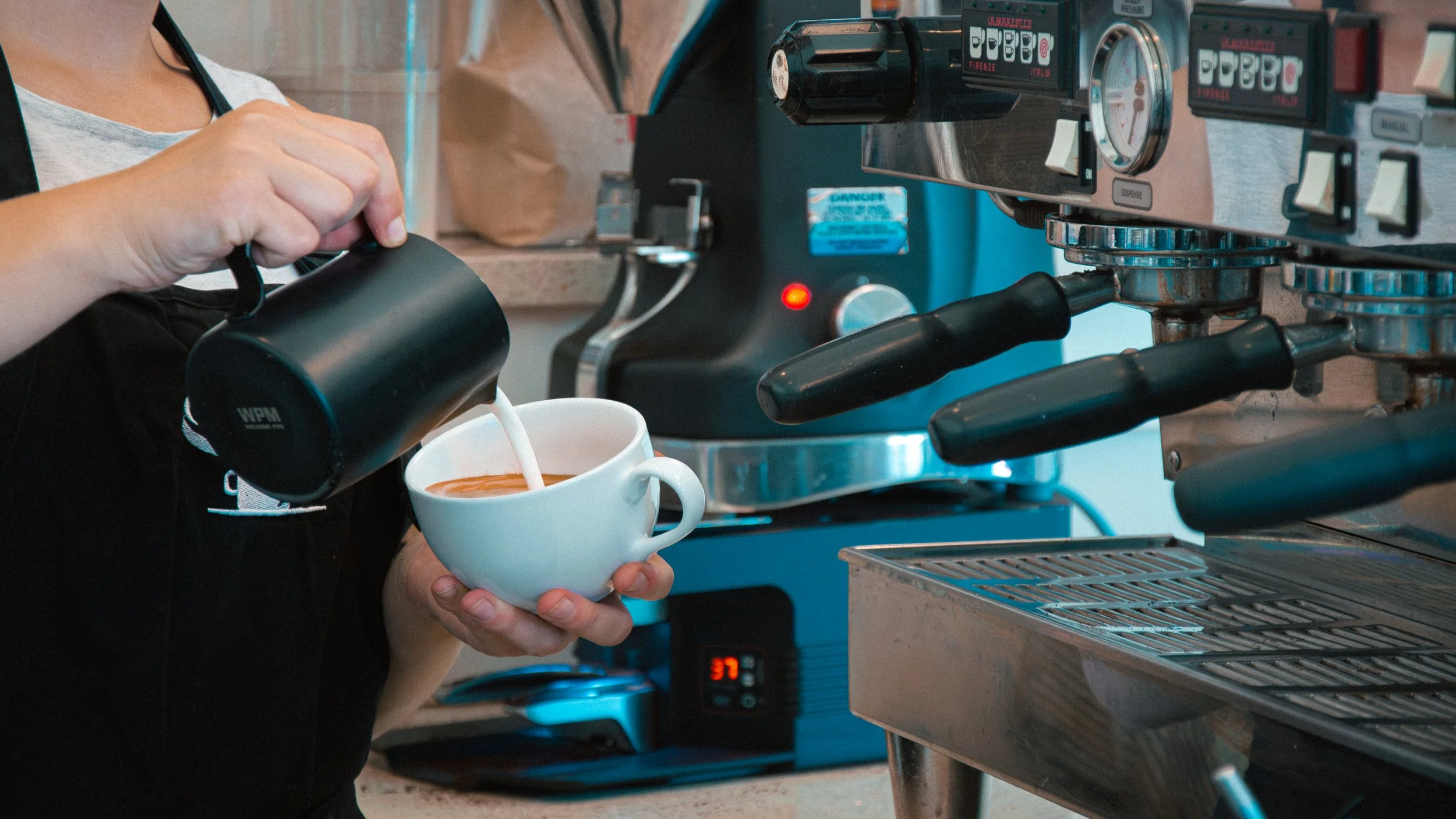Barista pouring steamed milk into a cup of espresso machine coffee at a coffee shop cafe.